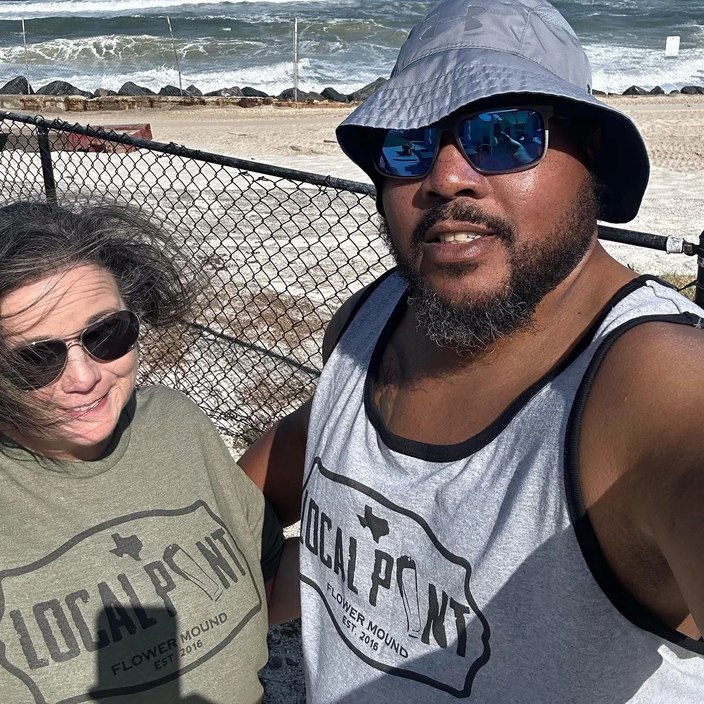Two people taking a selfie at the beach, standing in front of a chain-link fence with the ocean and rocks visible in the background. They are wearing sunglasses and casual tank tops.