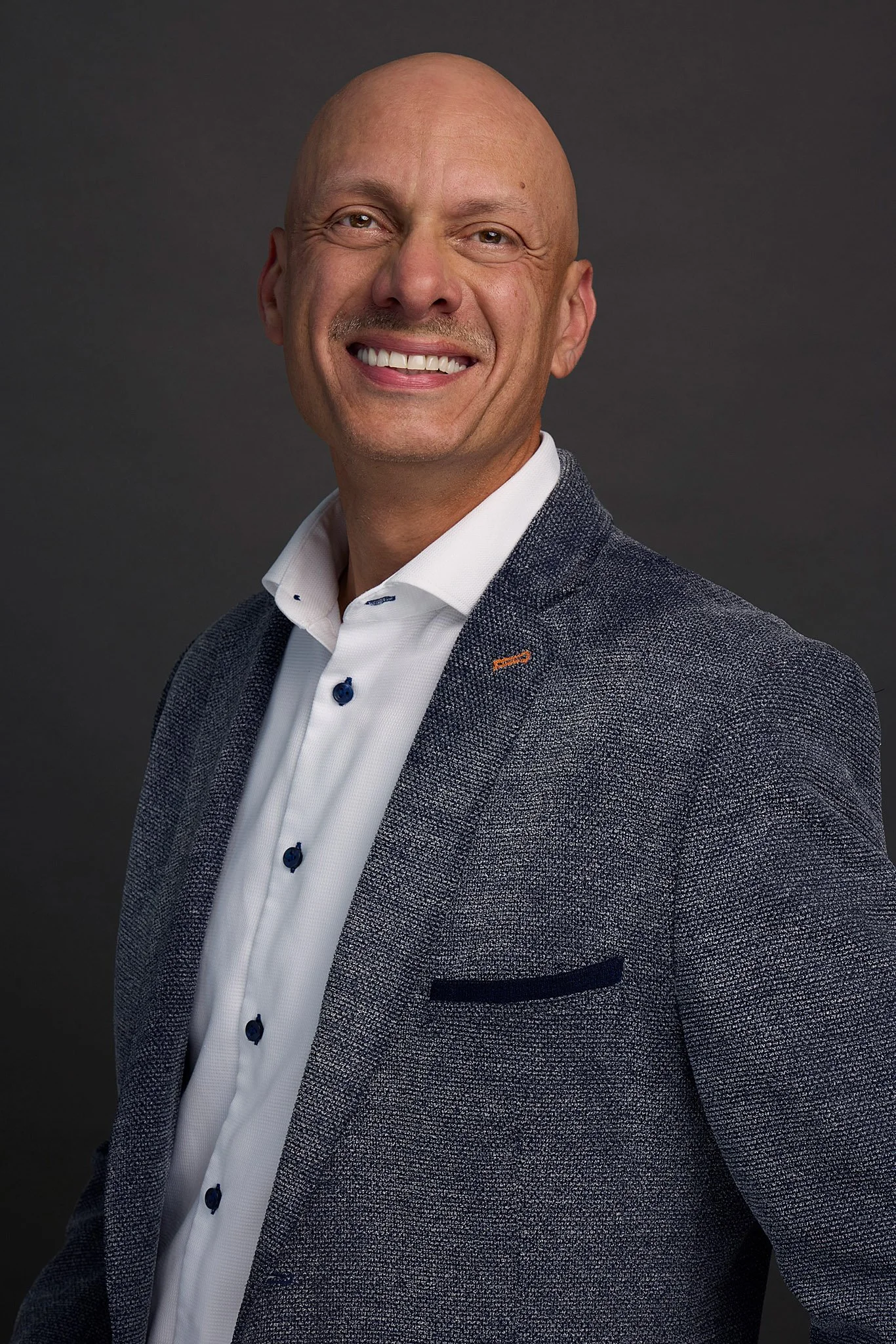 Tamer Mecky, a bald man with a warm smile, wearing a gray textured blazer over a white shirt, in a professional headshot against a dark backdrop.