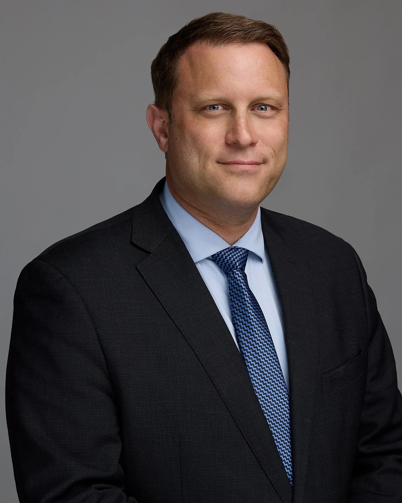 Devon Baily, a man with brown hair and blue eyes, smiling in a professional headshot against a gray backdrop.