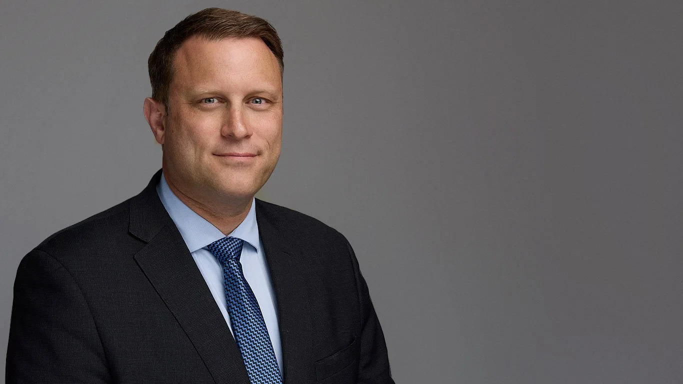 Devon Baily, a man with brown hair and blue eyes, wearing a suit and tie, in a professional headshot against a gray backdrop.