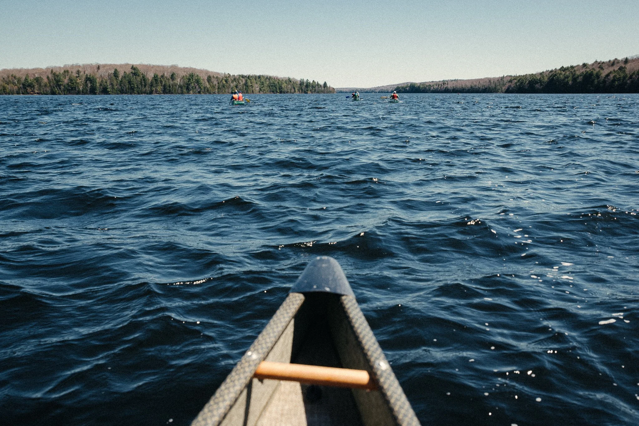 View from a canoe on a large lake with three kayakers in the distance and wooded shoreline in the background under a clear sky.