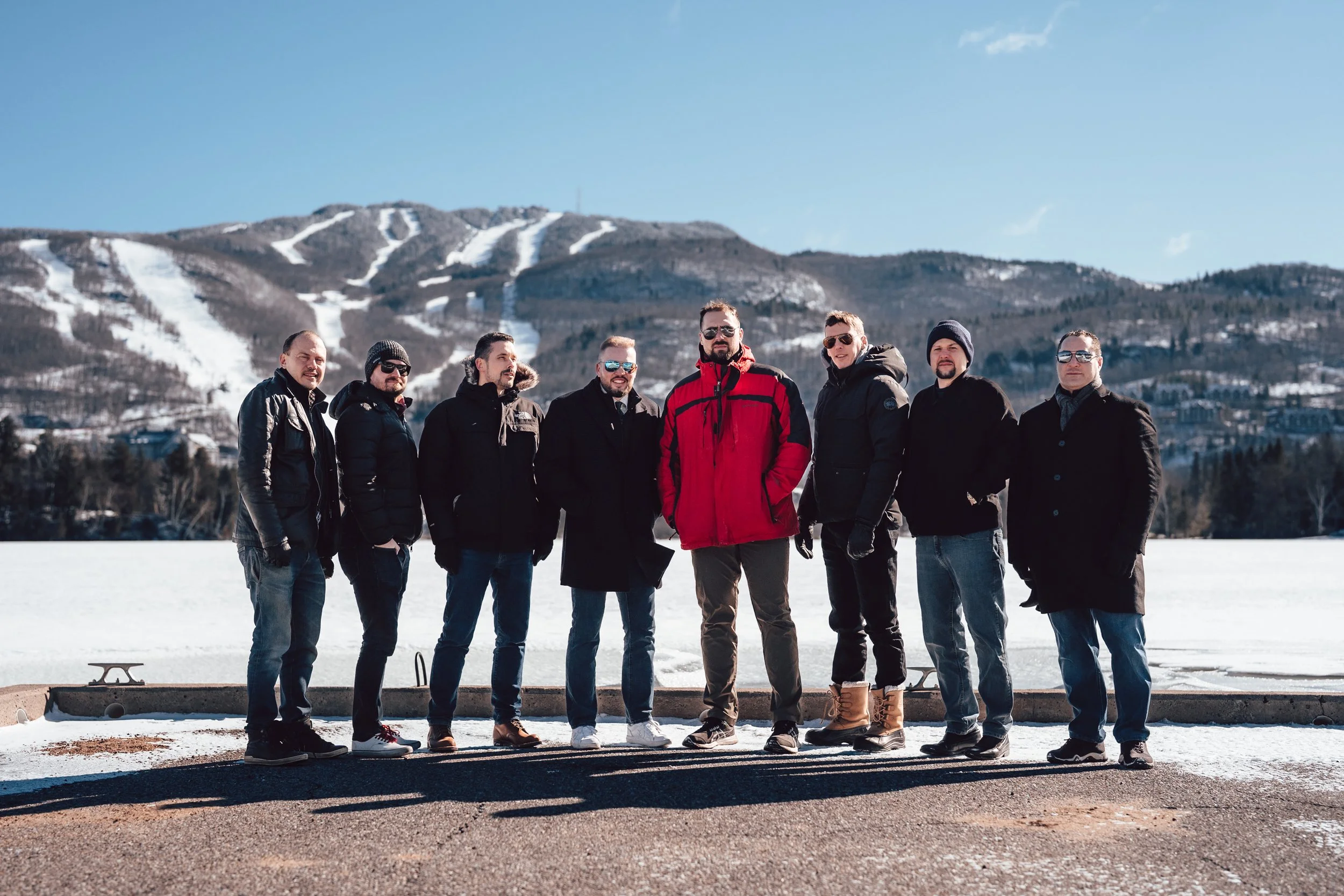 A group of eight men standing outdoors in winter clothing, with snow-covered mountains and ski slopes in the background.