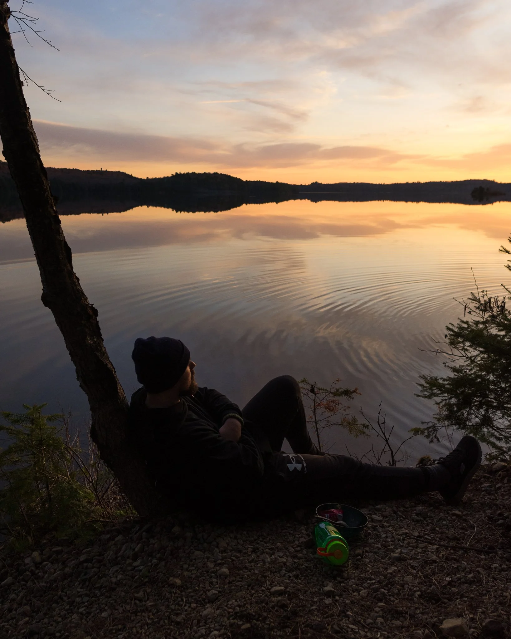 Person sitting by a lake at sunset, leaning against a tree, with headphones and a container nearby.
