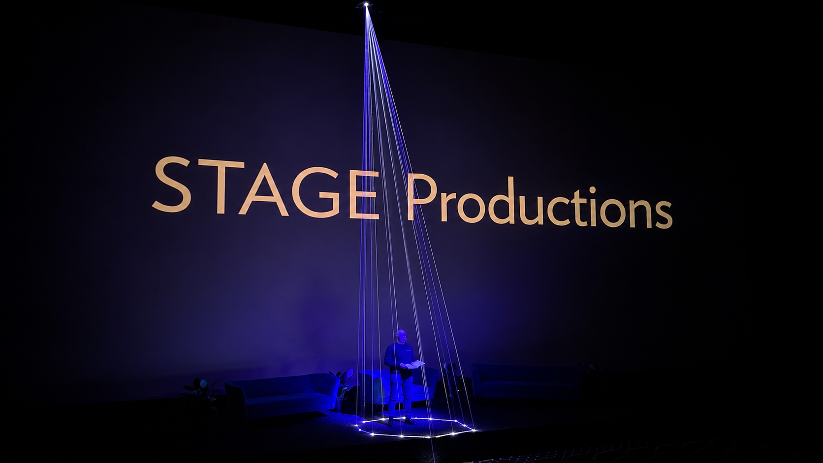 Stage with large screen displaying 'STAGE Productions', person standing on stage holding a book, illuminated by blue lights, and couches on either side of the stage.