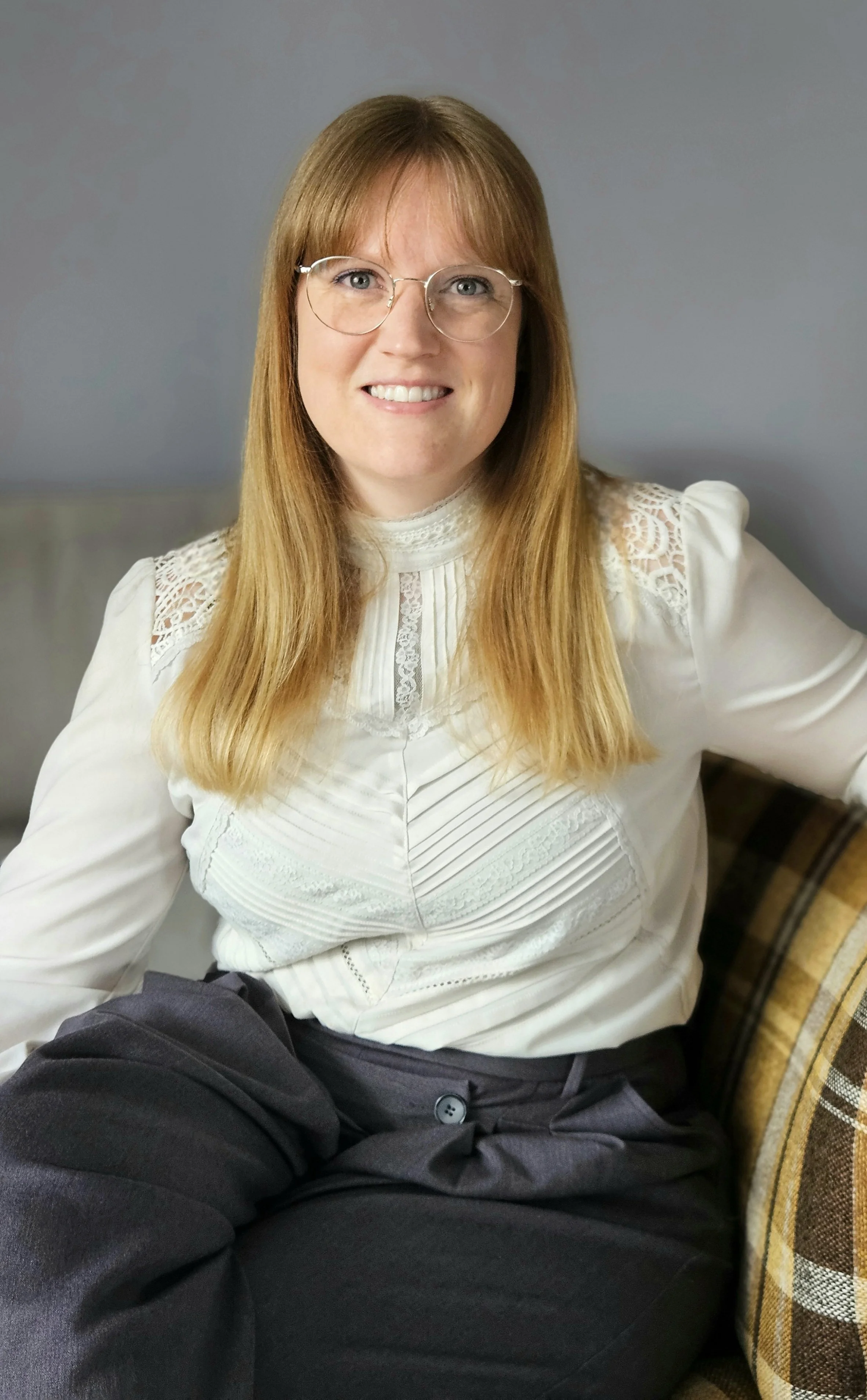 Eine lächelnde Frau mit Brille und roten Haaren, die in einem weißen Spitzenhemd sitzt, neben einem Mann. Das Bild ist in einem Raum mit grauem Hintergrund aufgenommen.