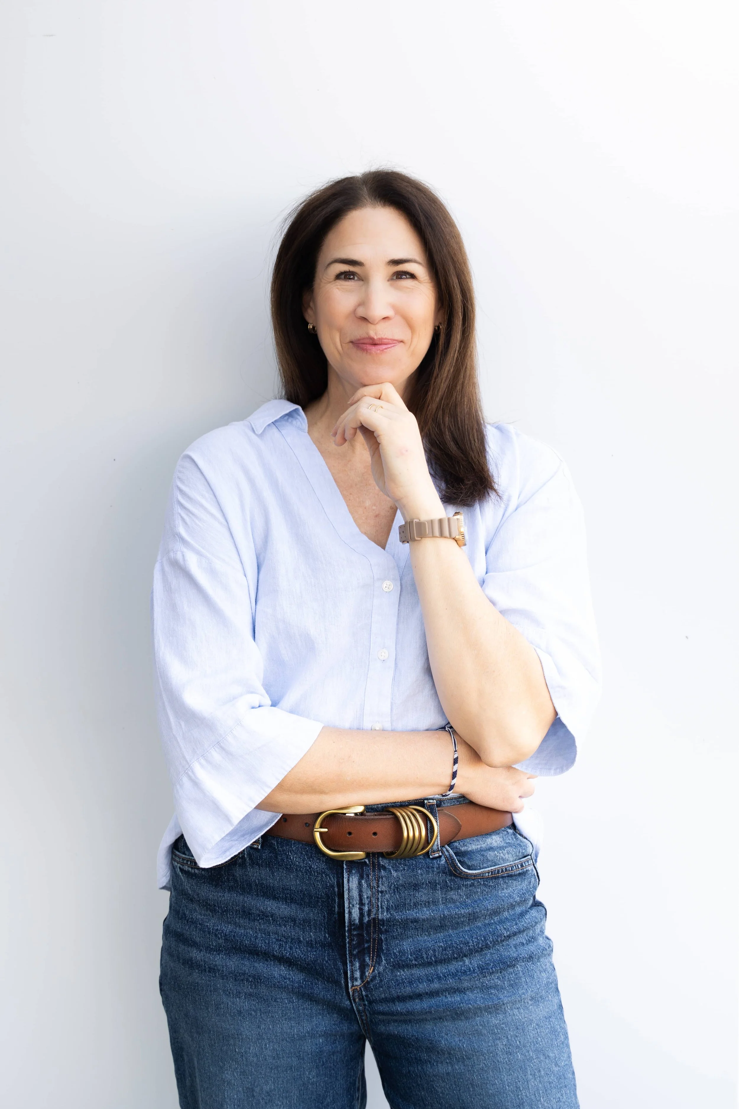A woman wearing a blue blouse and jeans, posing with her chin on her finger, smiling at the camera.