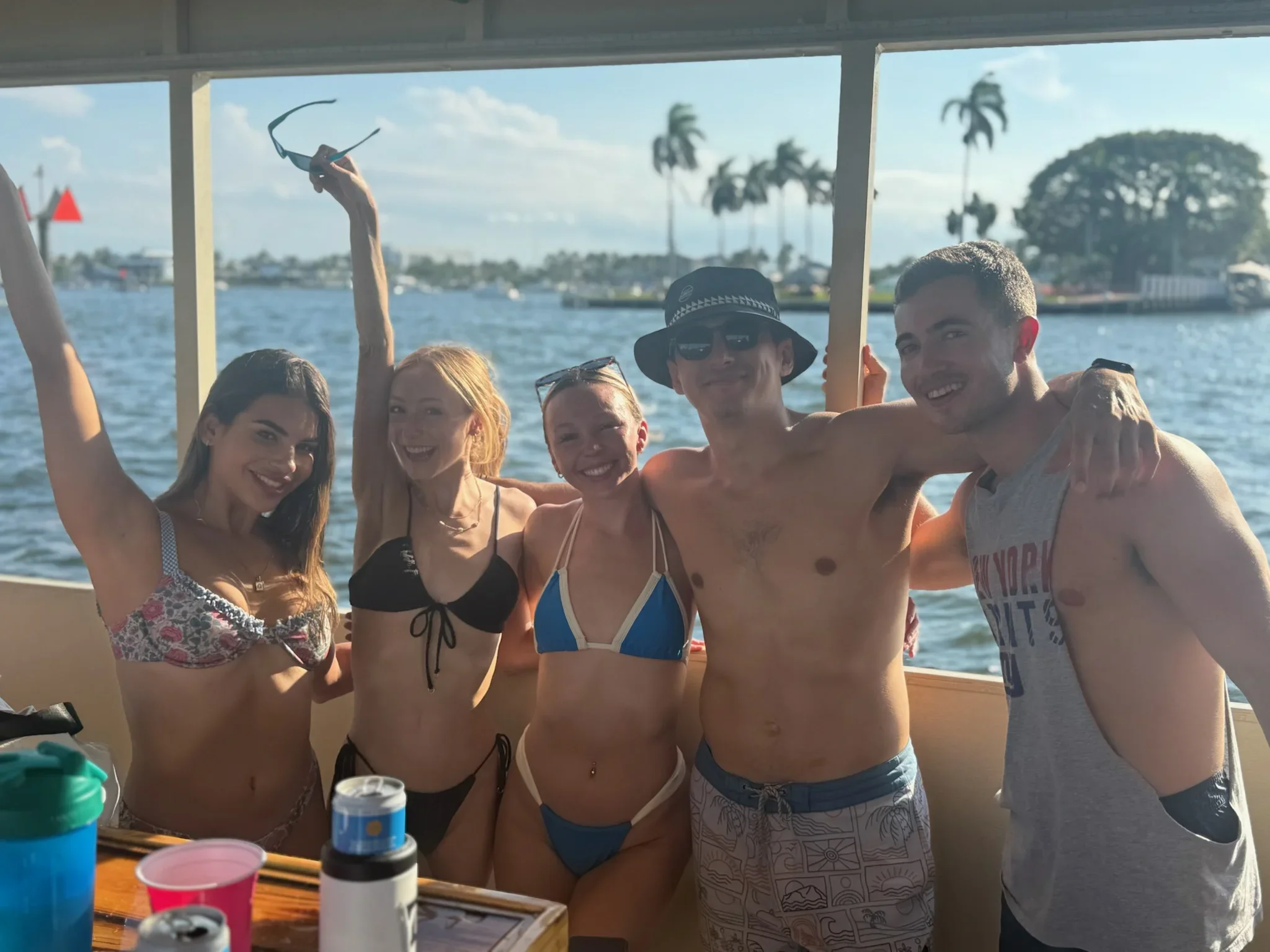 A group of young adults in swimsuits enjoying a boat ride on the Freaky tiki in Fort Lauderdale Florida, with palm trees and a blue sky in the background.