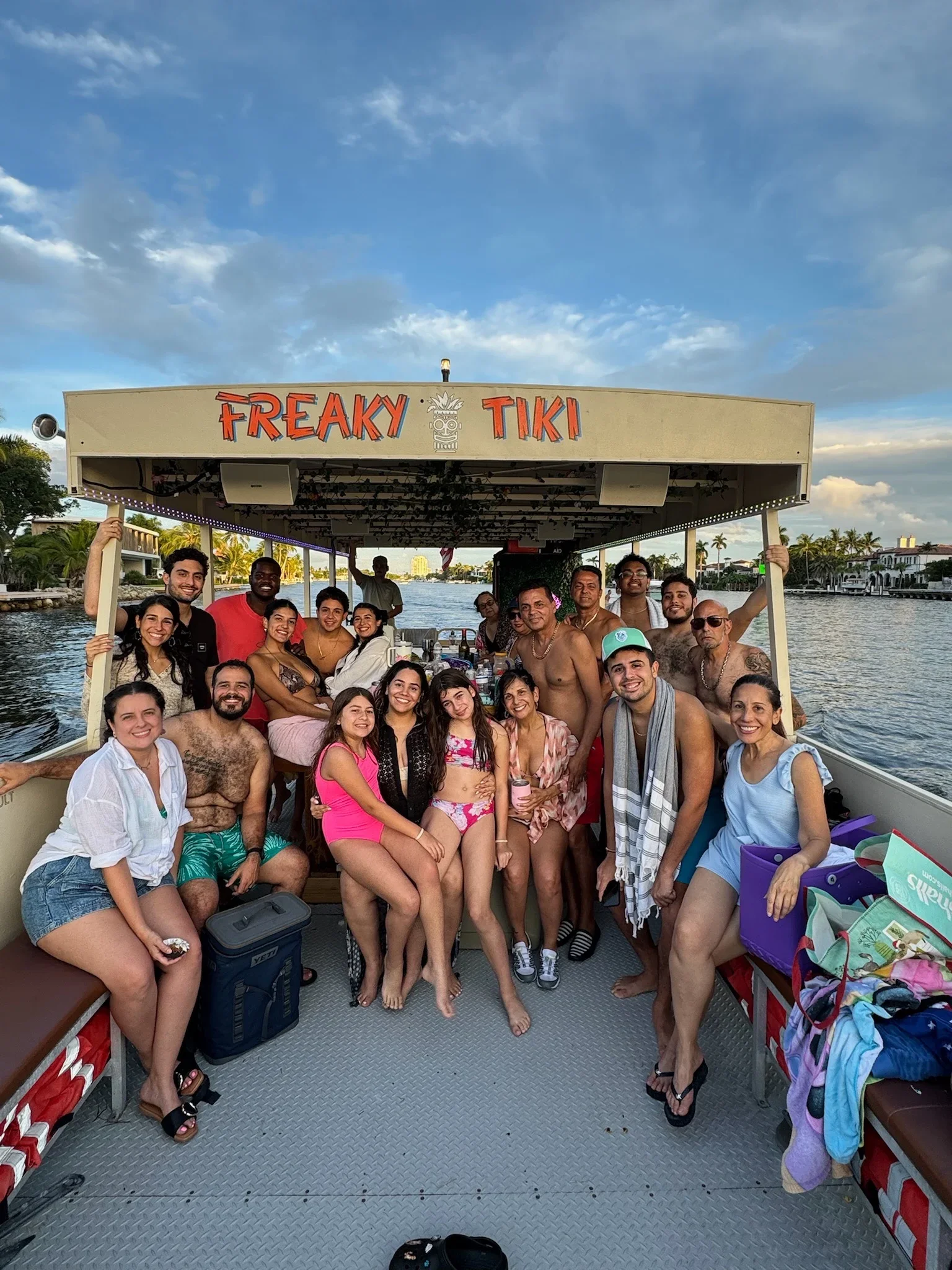 Group of people on a boat, posing and smiling, with water and houses with palm trees in the background. Freaky Tiki Fort Lauderdale Florida