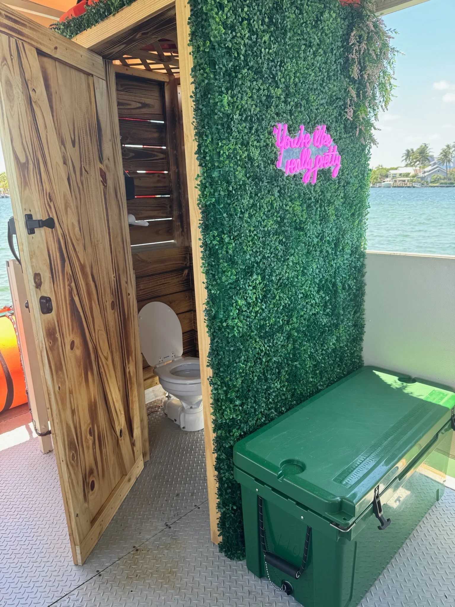 A wooden structure houses a toilet with a backdrop of a green hedge and a neon pink sign that reads 'You're the real pretty.' There is a green storage container in front of the hedge, Freaky Tiki Boat Charters Bathroom