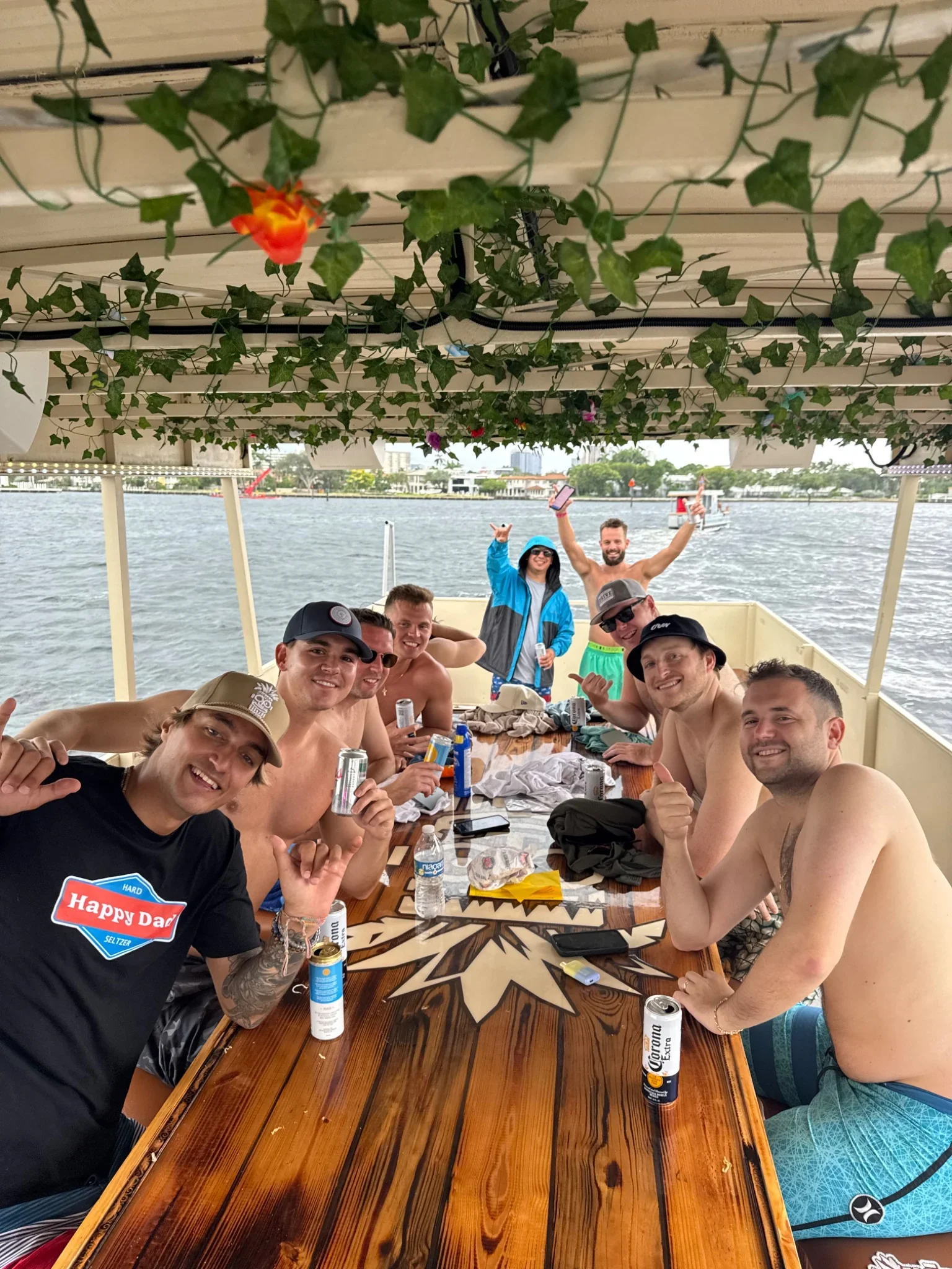 A group of shirtless men sitting around a wooden table on a boat The freaky tiki in fort lauderdale, smiling and raising drinks, with water and buildings in the background and rooftop greenery overhead.