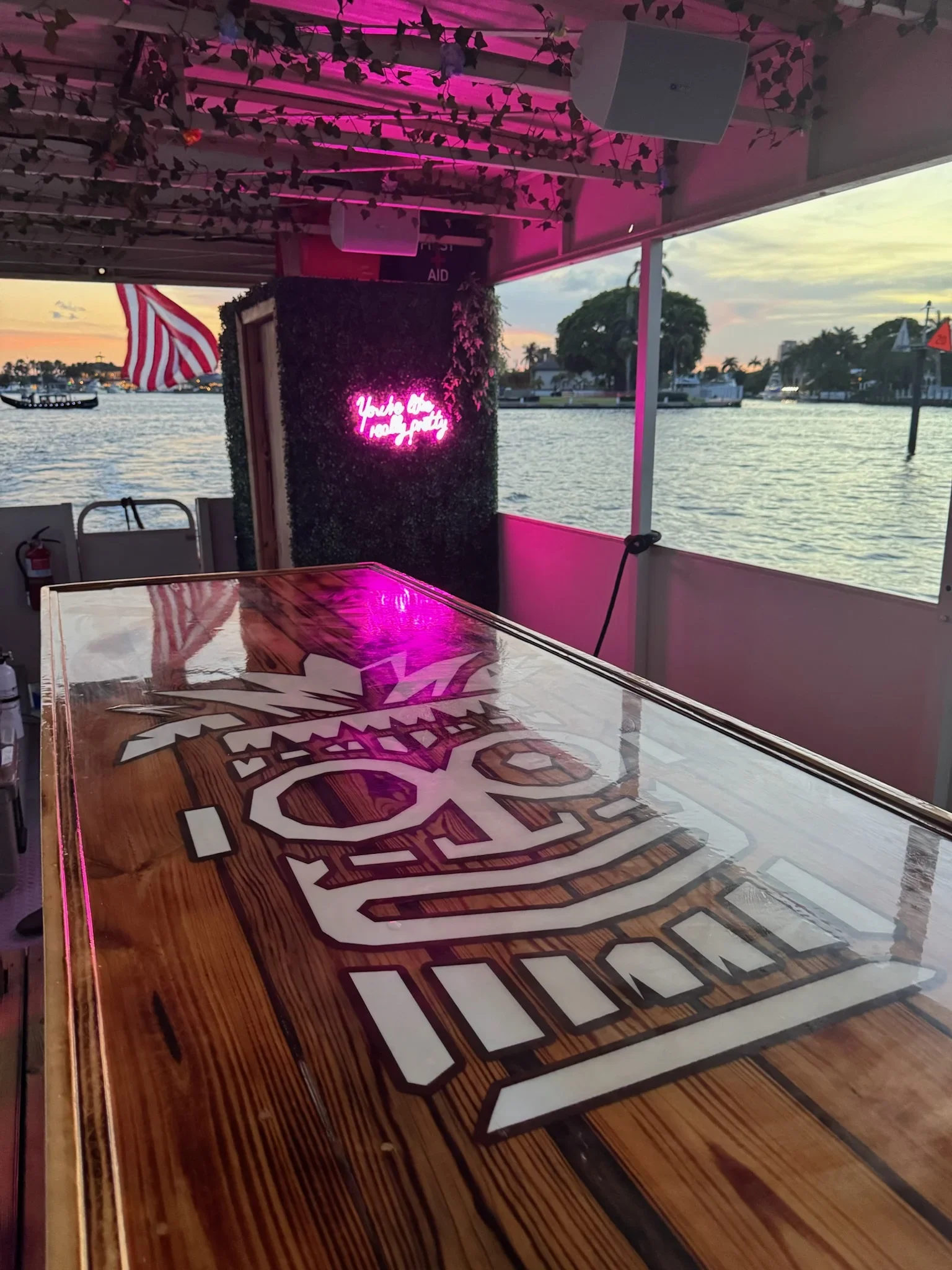 The image shows the interior of a boat or restaurant with a view of the water and boats outside. There is a neon pink sign that reads 'You’re the reason I stay' on a textured wall.  A wooden table with a logo of The freaky Tiki Fort Lauderdale