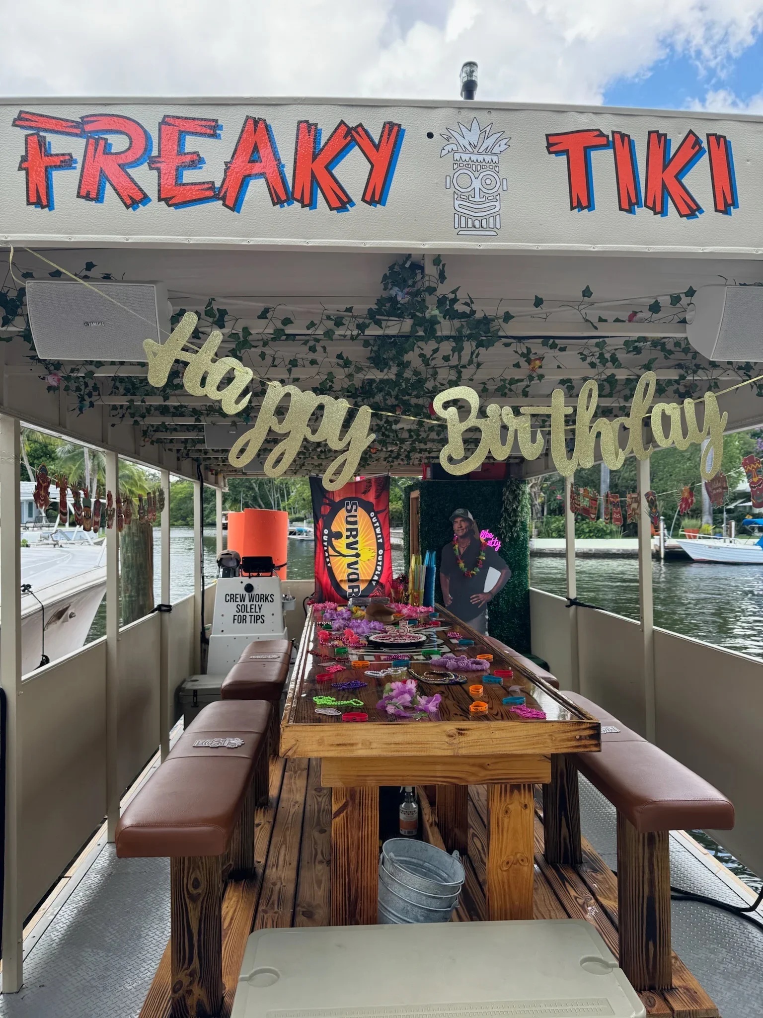 Inside a boat decorated for a birthday celebration with a “happy birthday” banner and various party accessories on a wooden table, overlooking a waterway with boats and trees. The Boat Says Freaky Tiki