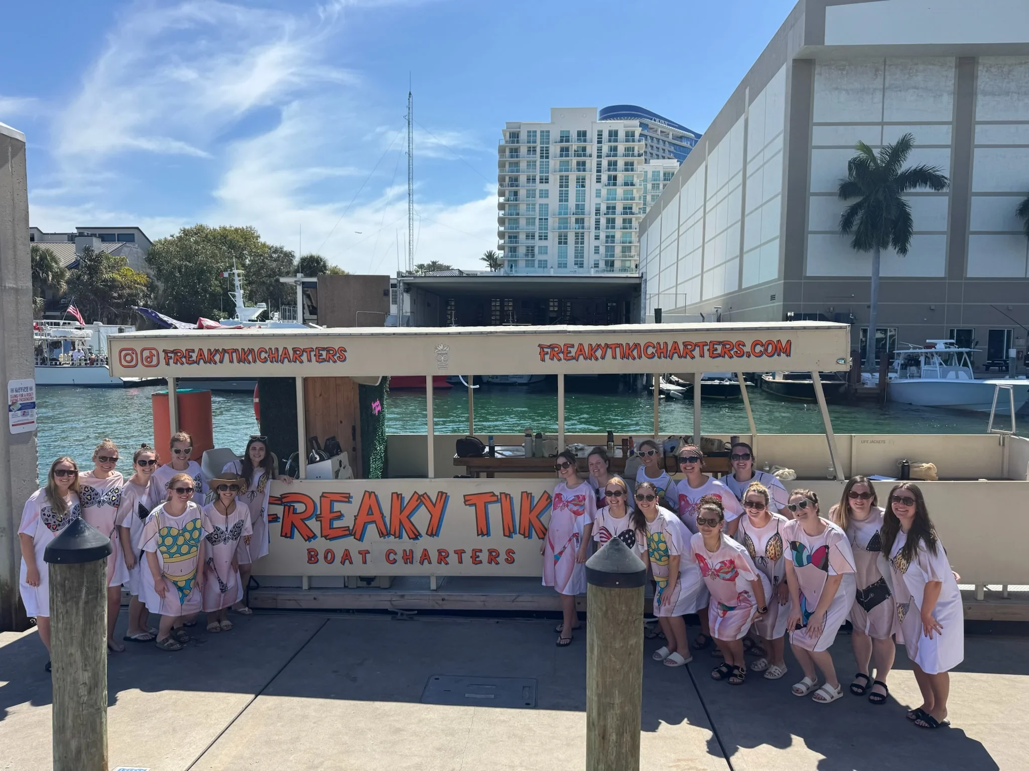 Group of people in matching white T-shirts with colorful designs standing in front of a boat charter sign that reads "Freaky Tiki Boat Charters" on a dock near water, with buildings and boats in the background.