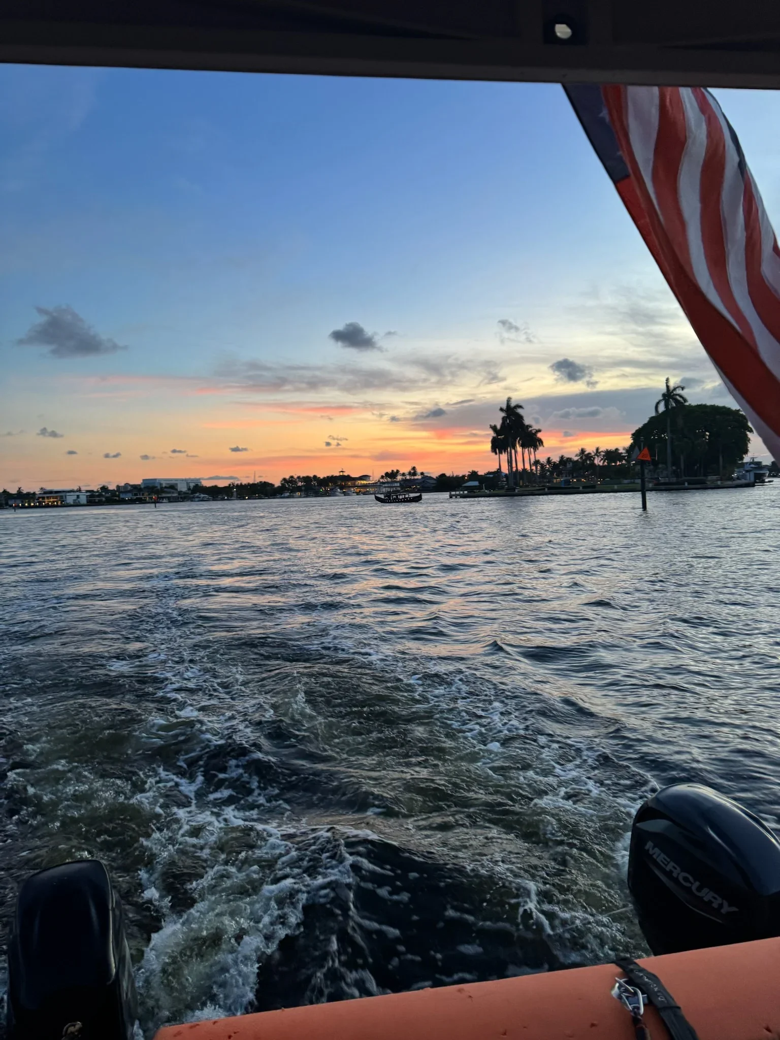 View from a boat on a waterway at sunset, with palm trees and buildings along the shoreline, and an American flag on the right side of the image. Freaky Tiki Boat Charters