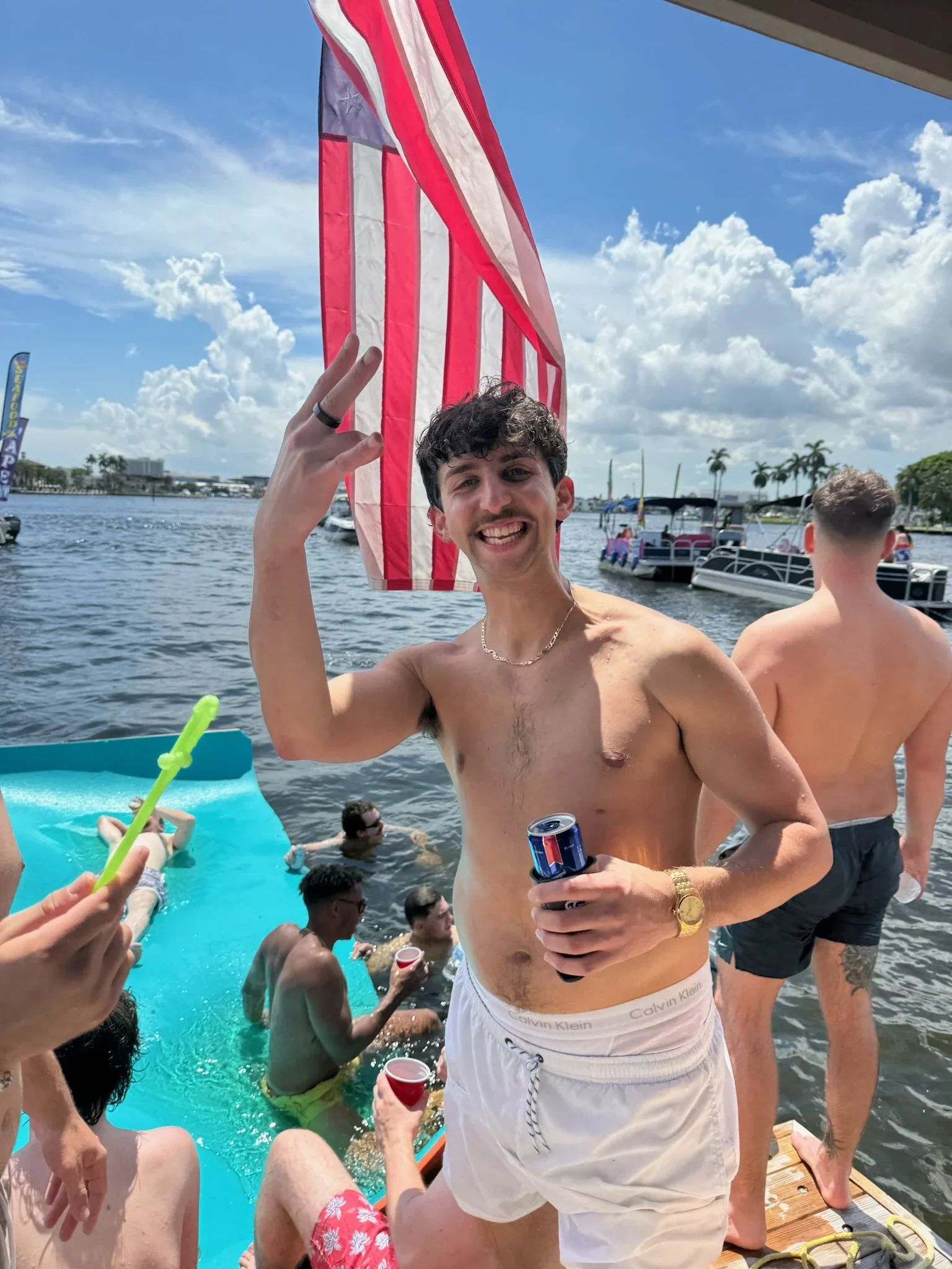 Young man with dark wavy hair, wearing white Calvin Klein underwear and shorts, smiling and holding a Red Bull can while standing on the freaky tiki in Fort Lauderdale Behind him, there are people swimming, boats, and a large American flag.