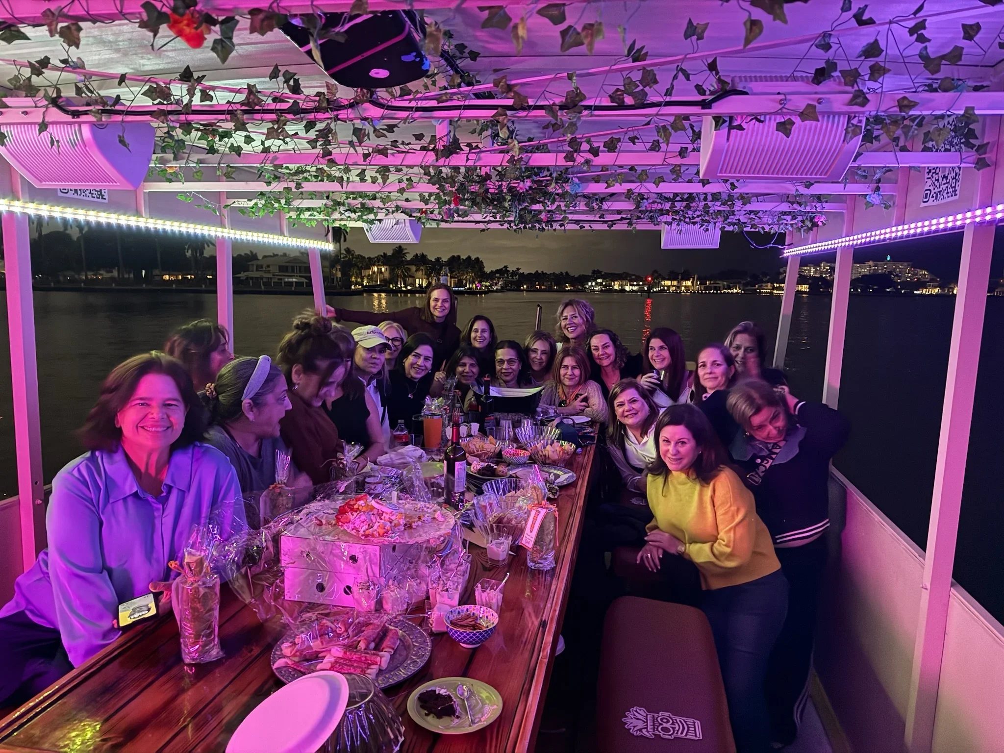 A group of women gathered around a dining table on the freaky Tiki at night, smiling for the camera. The boat is decorated with purple and pink lights, and there are food and drinks on the table. The background shows water and distant buildings.
