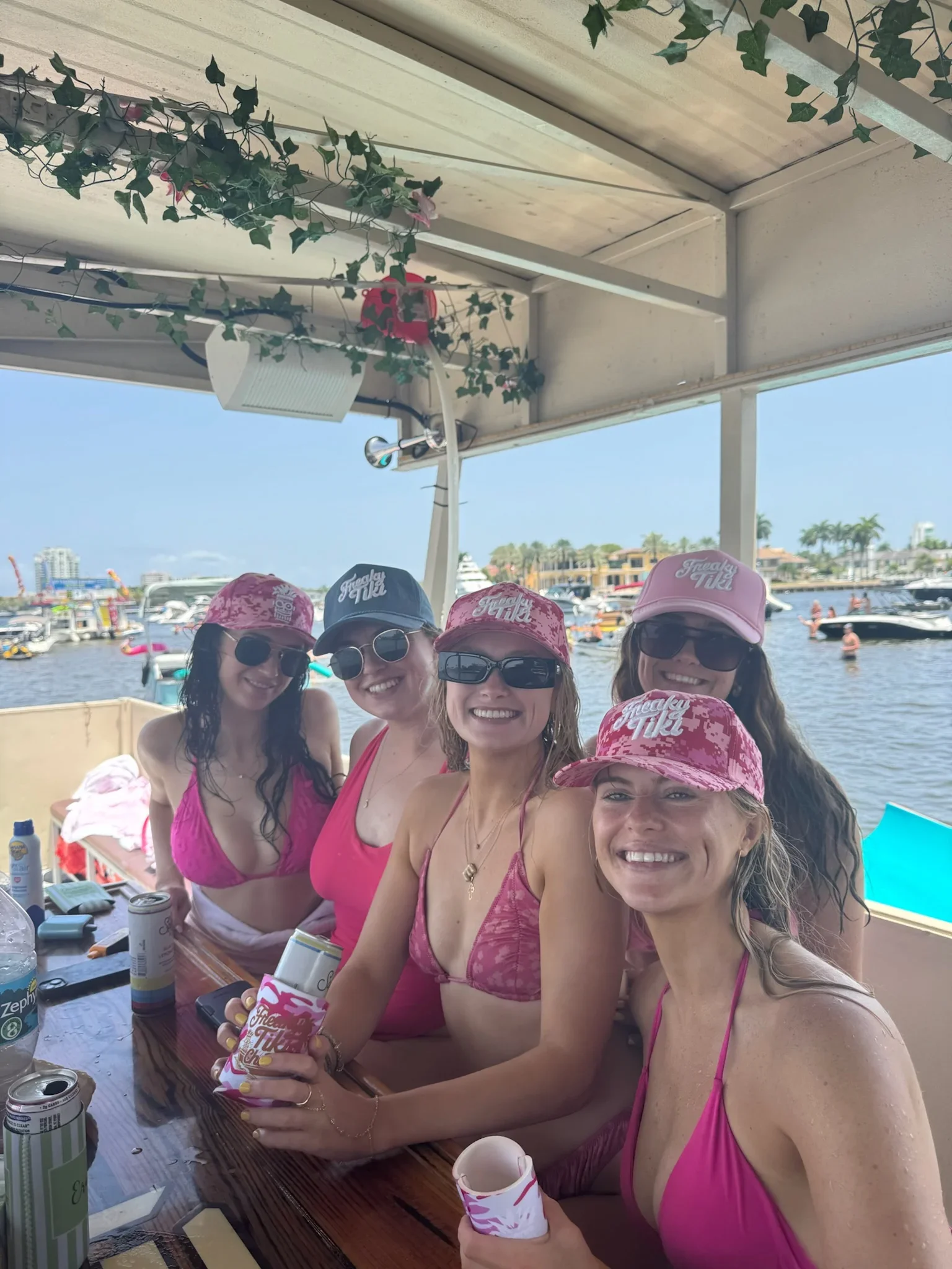 A group of women wearing pink swimsuits and pink hats with 'Freaky Tiki' written on them, sitting at a table outdoors near water, smiling, with boats and boats in the background. Freaky Tiki Boat Charters Fort Lauderdale