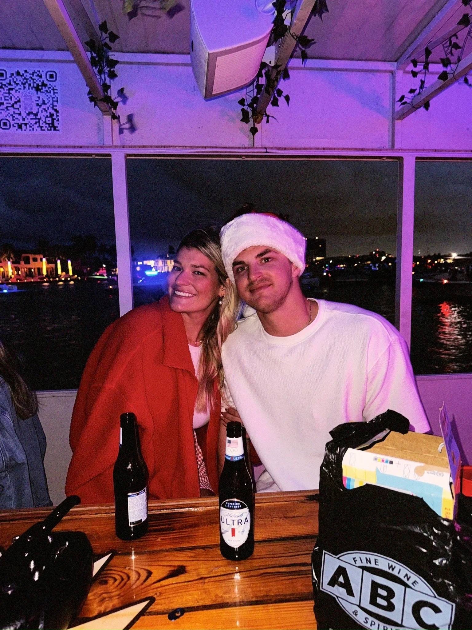 A man and woman sitting at a table on the freaky tiki boat at night,t he woman wears a red jacket and the man wears a Santa hat. There are two beer bottles on the table, and a bag of ABC wine. The background shows city fort Lauderdale