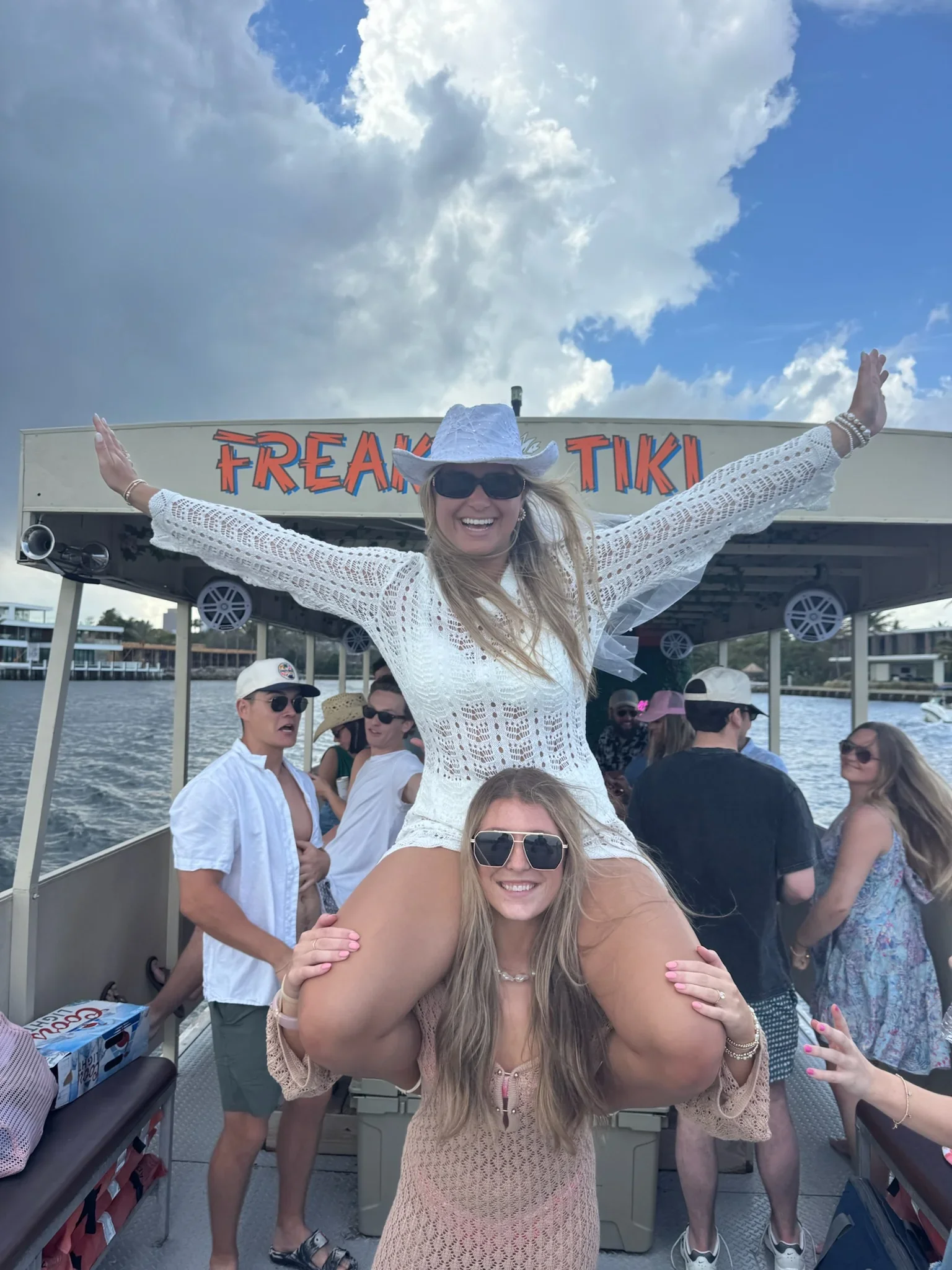 People enjoying a boat ride on the freaky tiki in Fort Lauderdale its a sunny day, with the woman at the top wearing a white cowboy hat, sunglasses, and a white lace dress, smiling with arms outstretched.