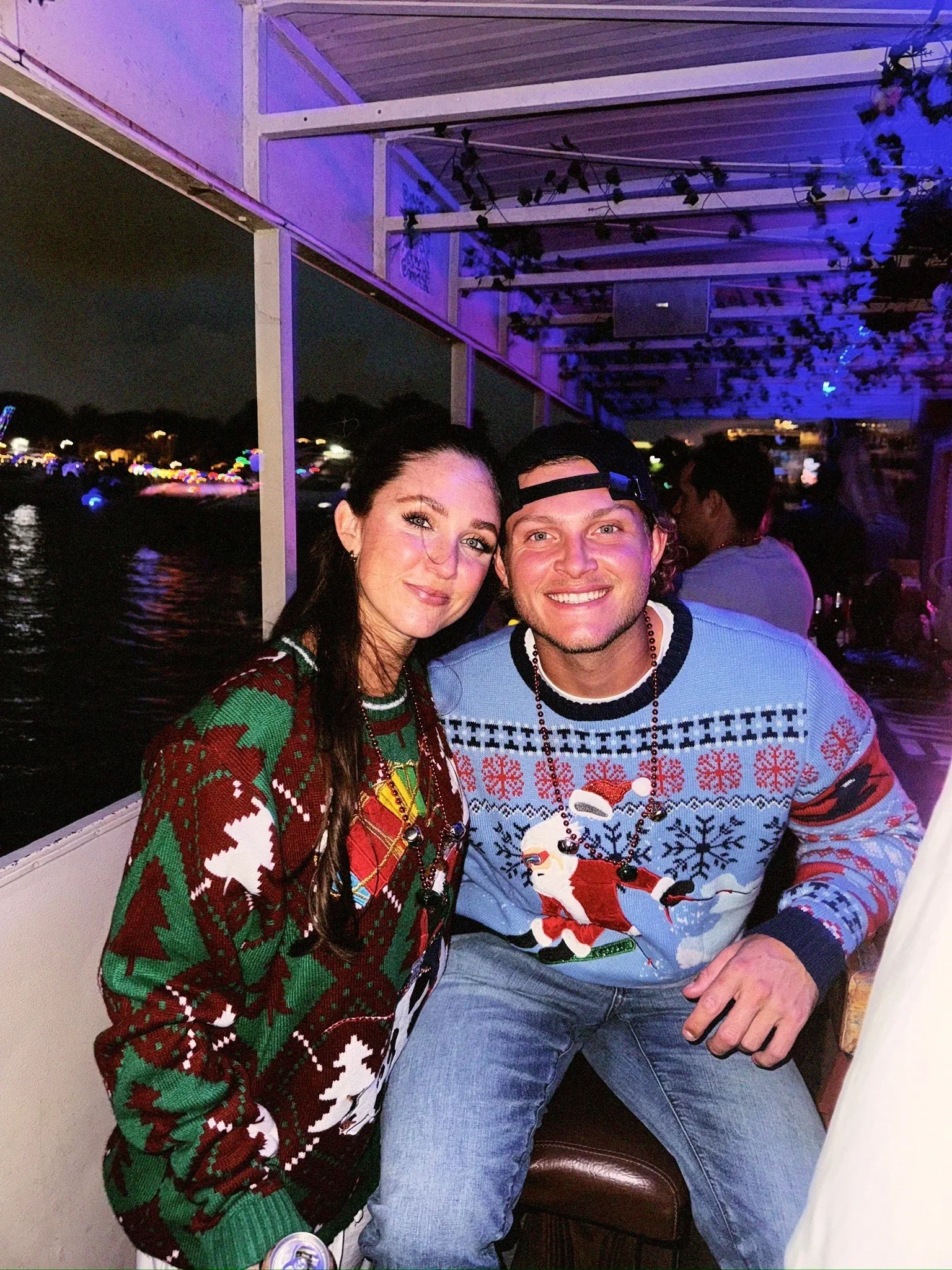 A man and woman sitting together on a boat at night, both smiling. They are wearing Christmas sweaters with festive patterns. The night sky and waterfront with colorful lights are visible in the background. Freaky Tiki Christmas Party