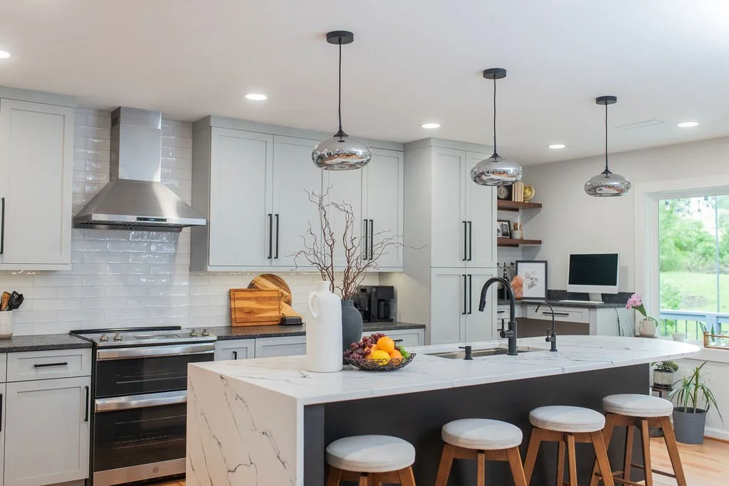Modern kitchen with gray cabinets, stainless steel stove and range hood, white subway tile backsplash, marble island with bar stools, pendant lights, and a small workspace with a computer near a large window.