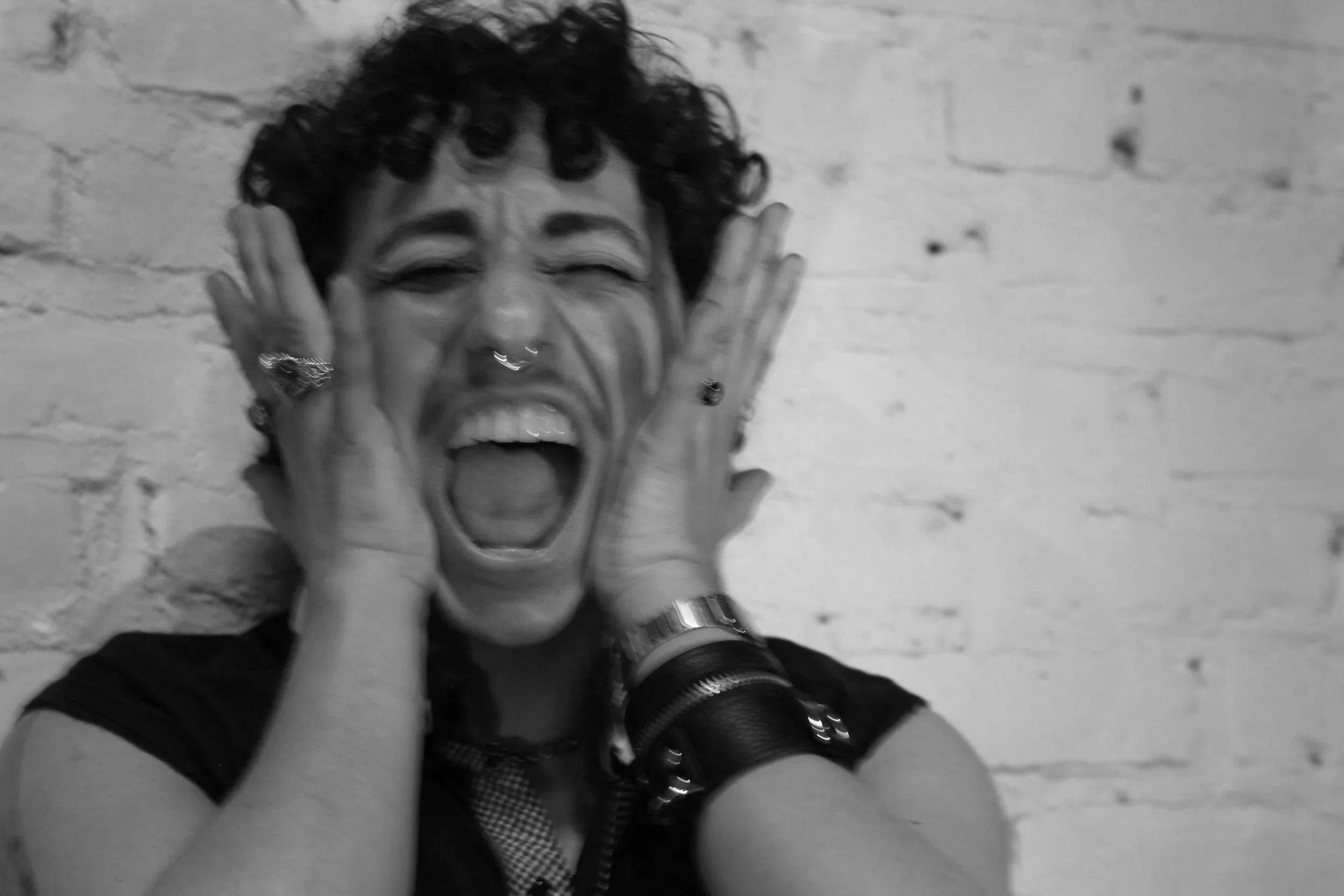 A person with short curly hair and multiple rings on his fingers holding his face and screaming. He is wearing multiple jewelry items on his hands and a dark top, against a light-colored brick wall background.