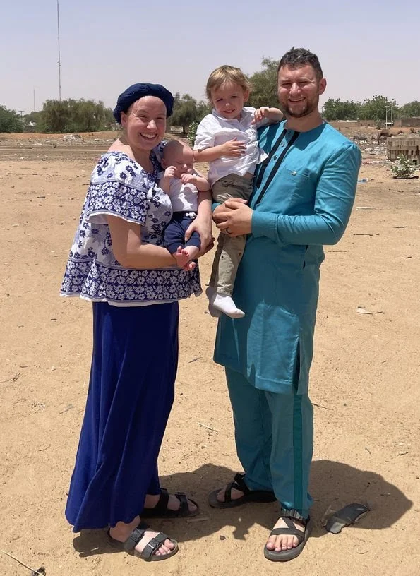 A happy family of four standing outdoors in a desert-like area, with two women holding two young children, all smiling at the camera.
