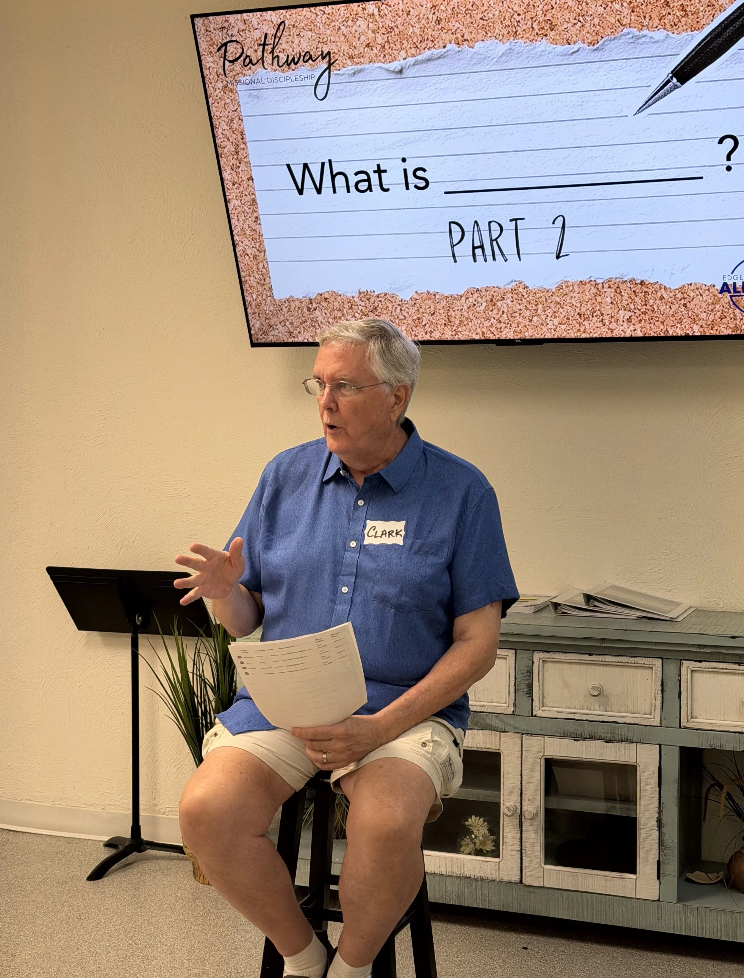 A man with gray hair wearing glasses, a blue shirt, and shorts is sitting on a black stool. He is holding papers and appears to be speaking. Behind him, there is a wall-mounted screen displaying a presentation slide titled 'The Pathway' with the subtitle 'MISSIONAL DISCIPLESHIP' and the question 'What is ___ ? PART 2'. There is also a black music stand, a potted plant near the stool, and a distressed white cabinet with drawers in the background.