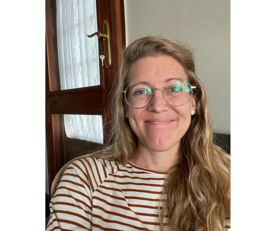A woman with long wavy blonde hair wearing glasses and a striped shirt, smiling at the camera inside a house near a window with curtains.