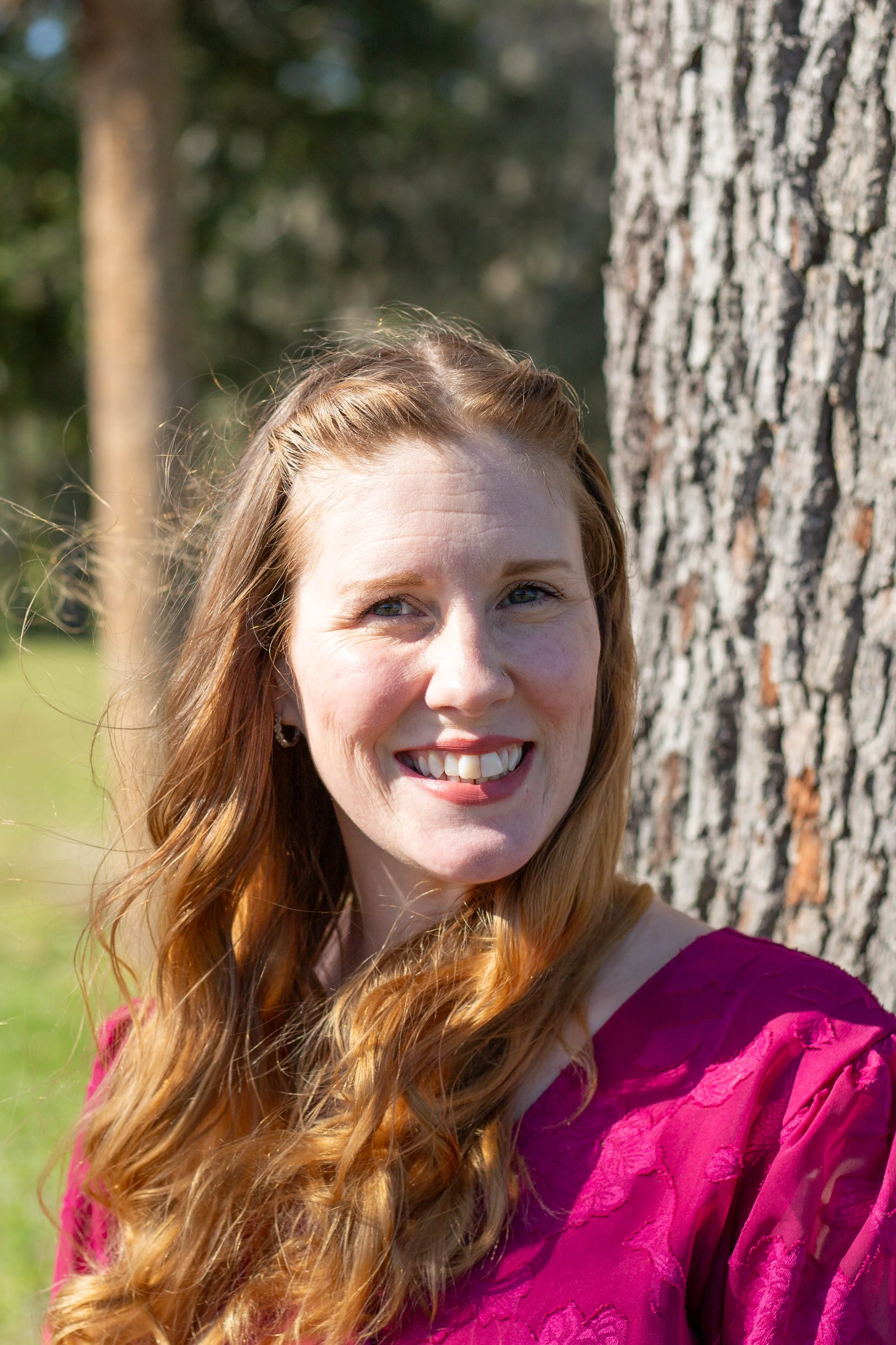 A woman smiling outdoors with trees in the background. She has red hair and is wearing a magenta dress with floral lace patterns.