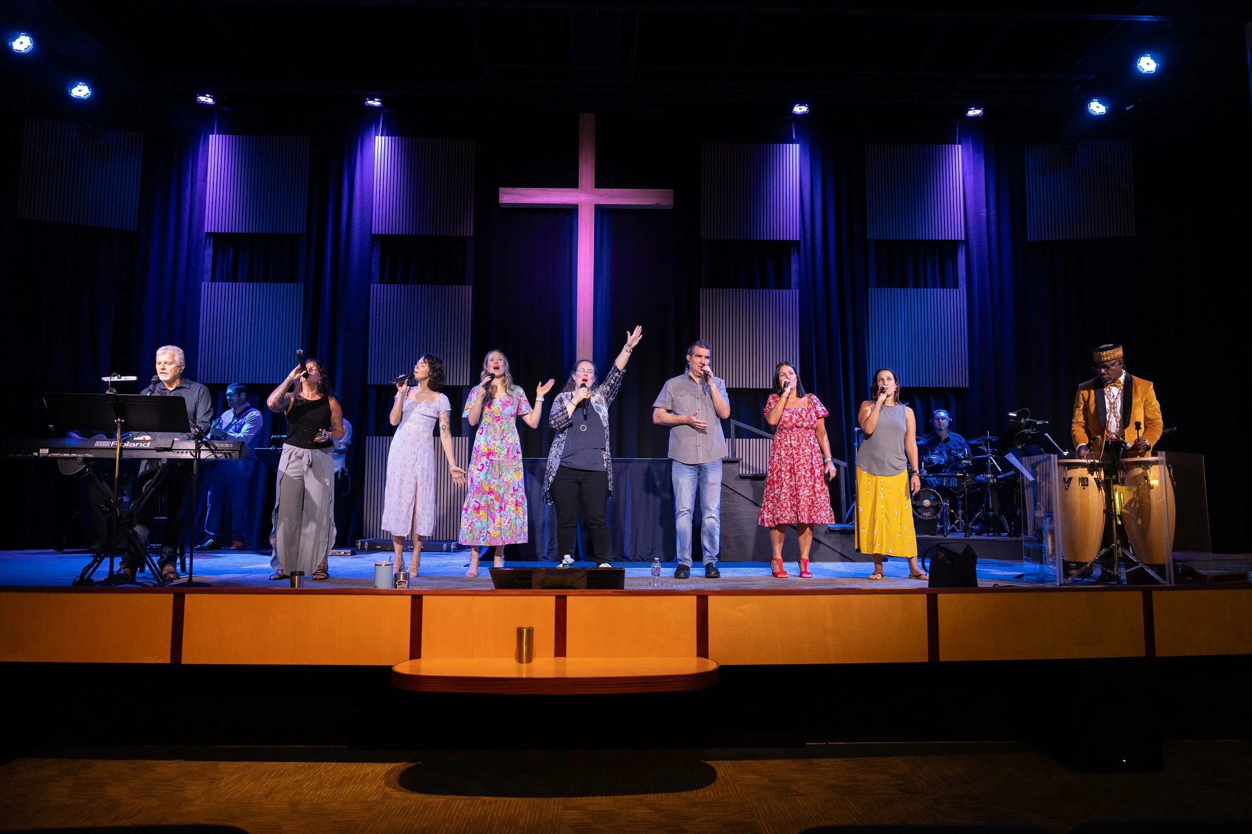 A group of five singers performing on stage in a church with a large illuminated cross in the background, accompanied by a musician playing conga drums.
