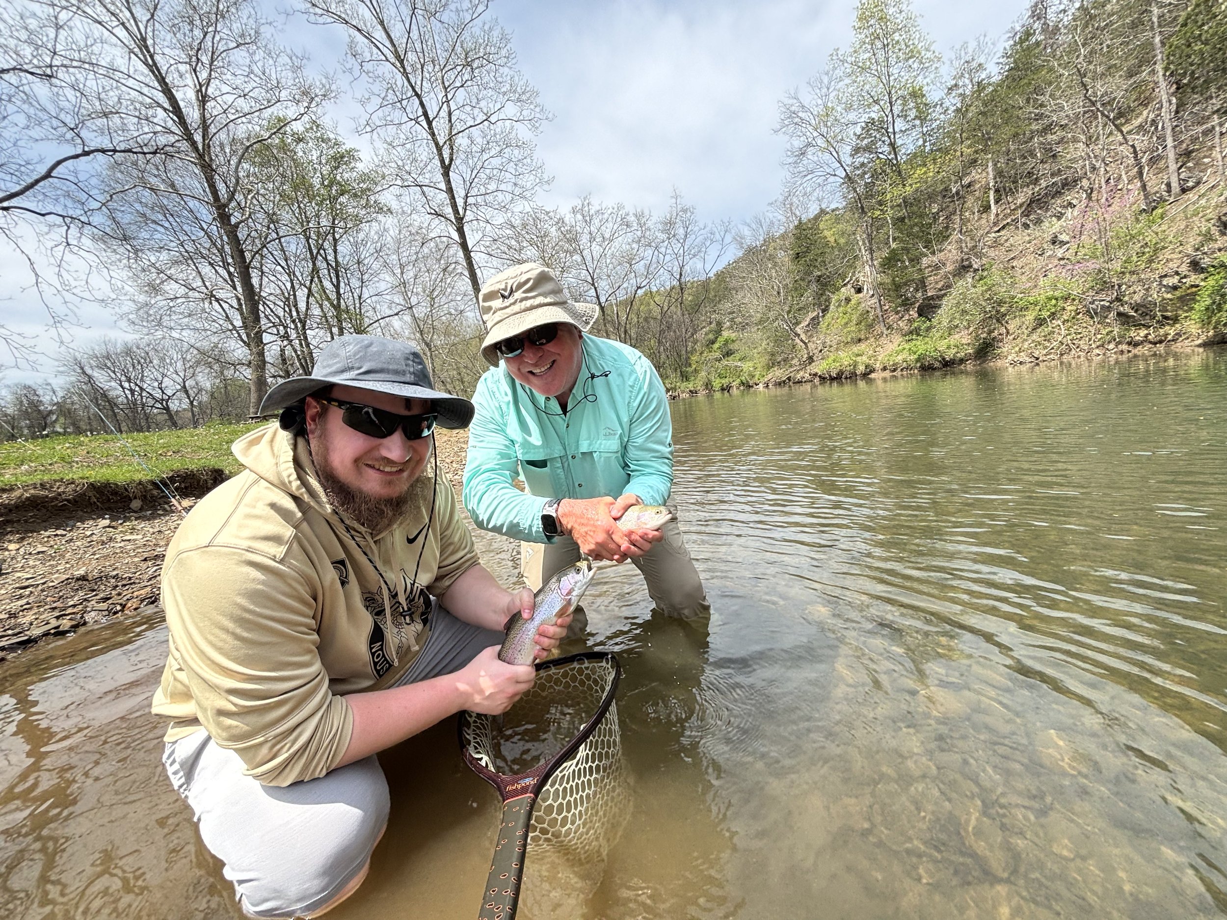 Two men fishing in a river, smiling and holding a fish above the water, with trees and a rocky bank in the background.