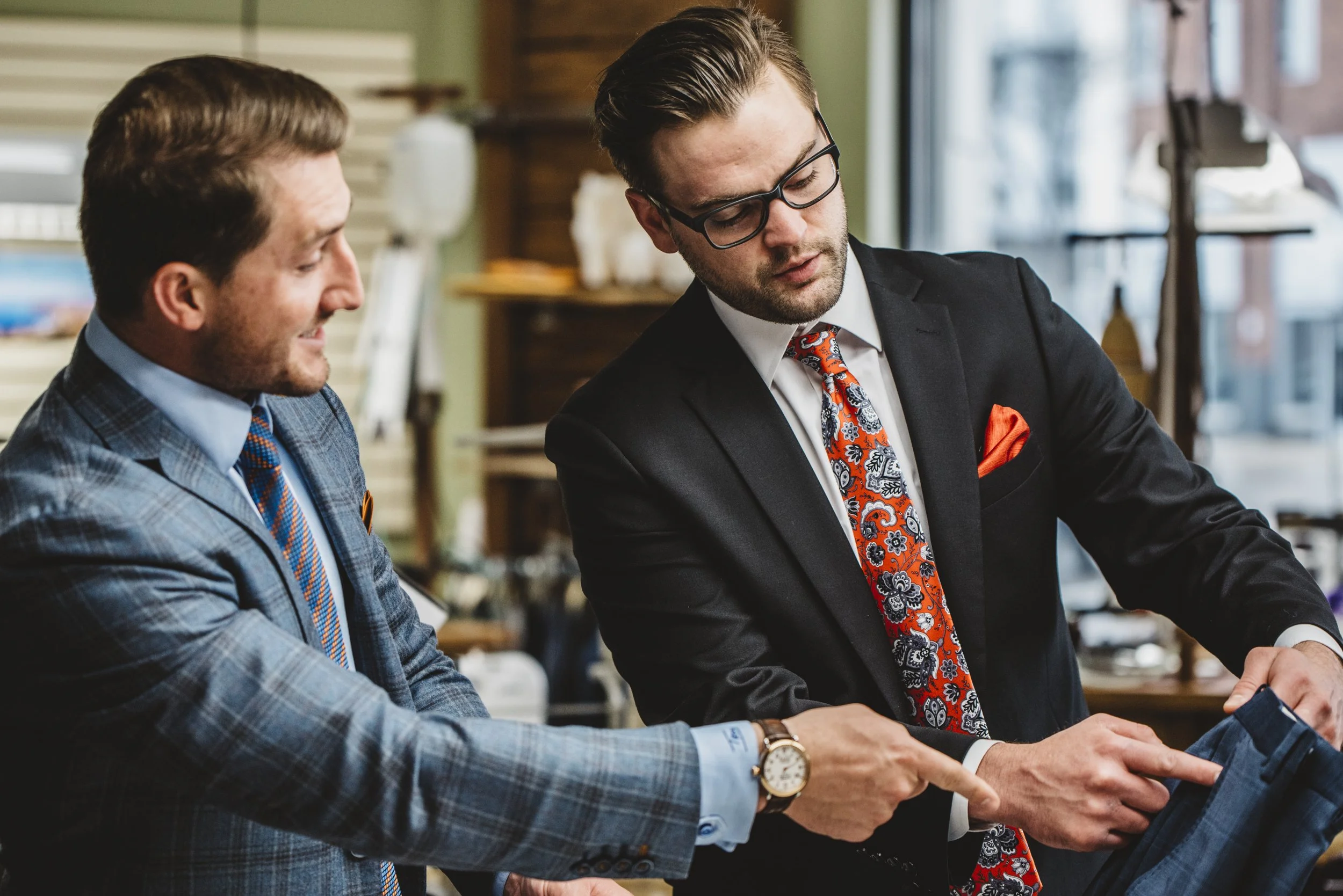 Two men in suits and ties standing and looking at a pair of pants in a boutique.