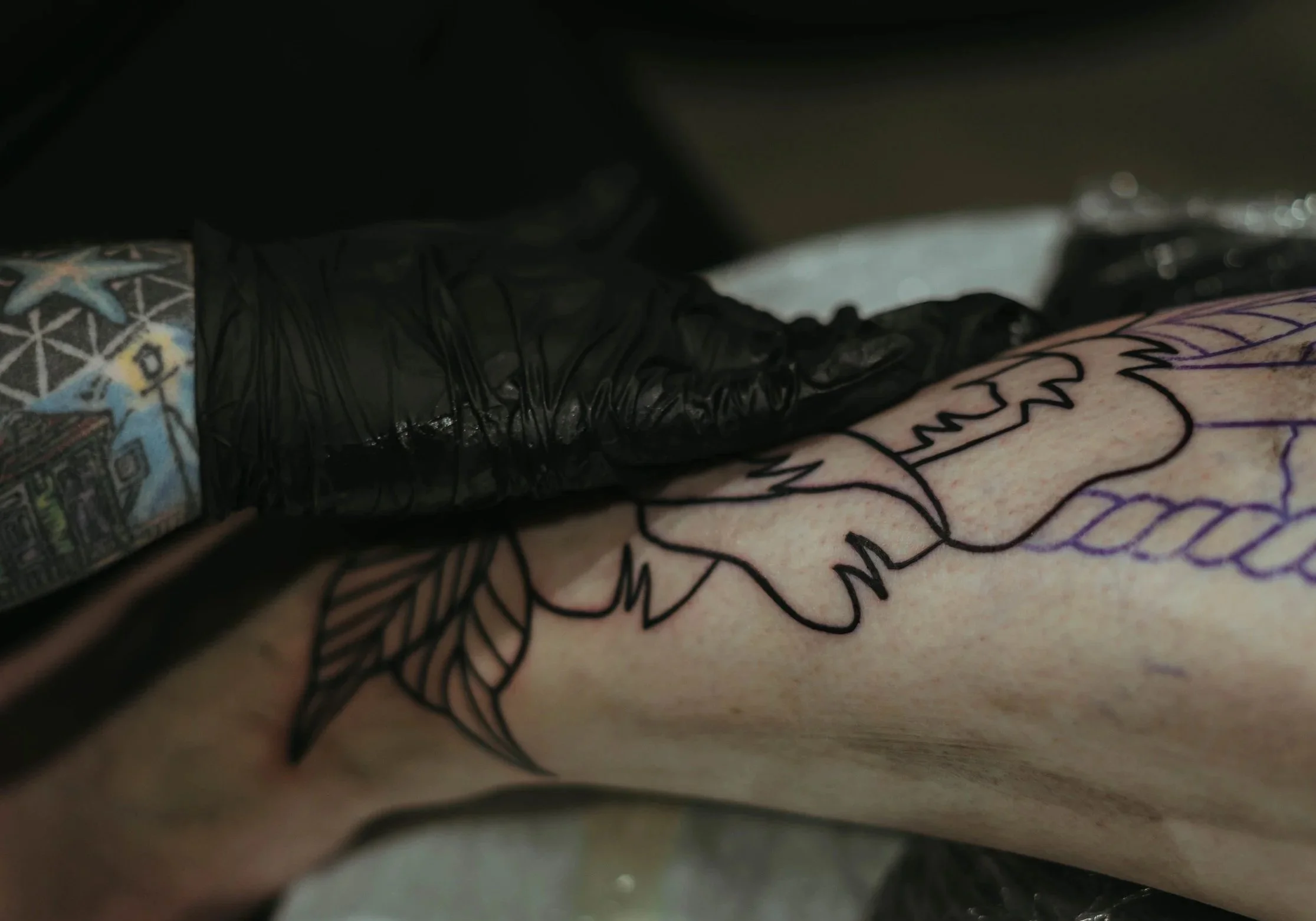 Close-up of a tattoo on a person's skin, with colorful ink and black outlines, partially obscured by black plastic and shadow.