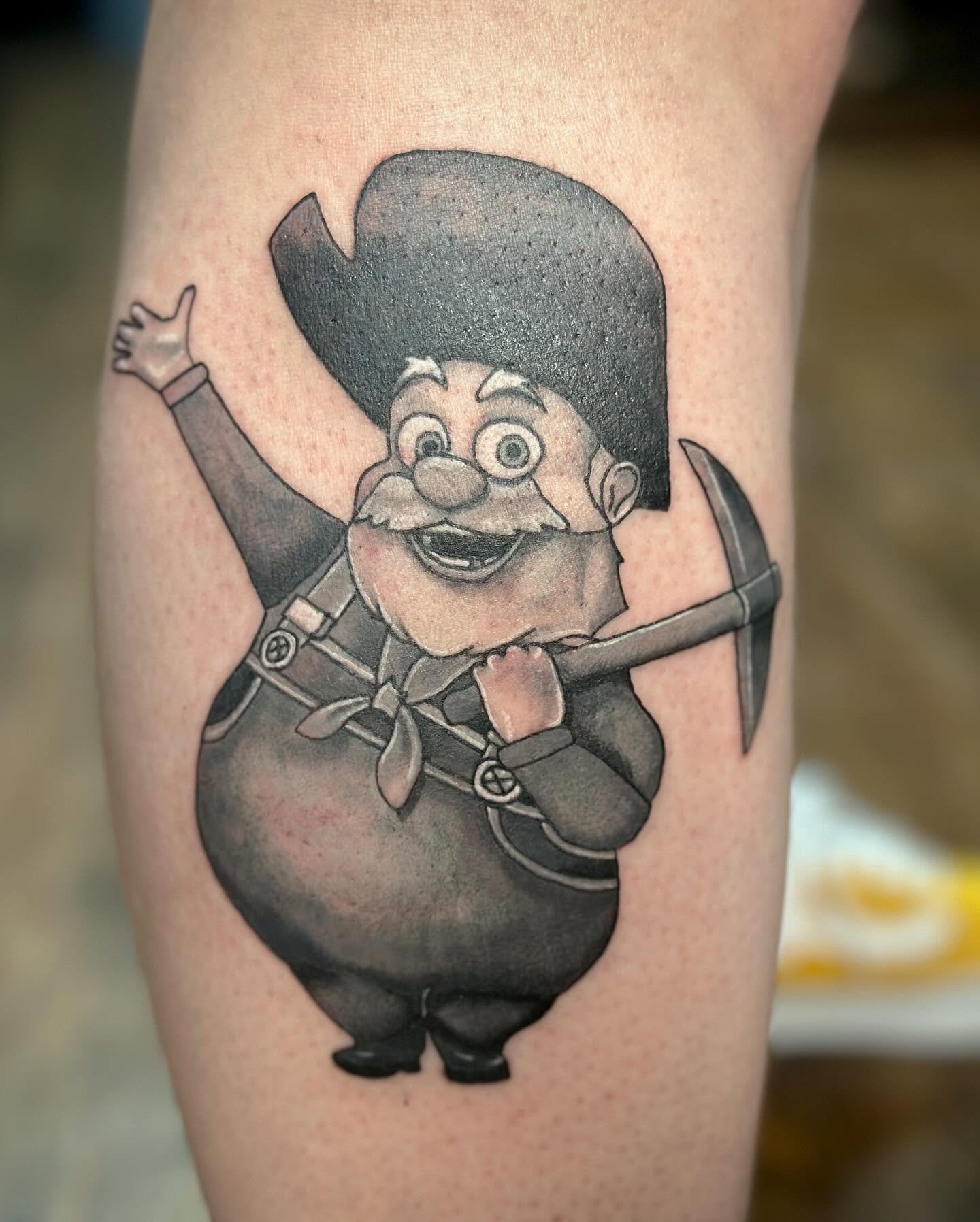 Tattoo of a cartoon dwarf with a big hat, mustache, and a broad smile, dressed in a dark outfit with a scarf, holding a sword with both hands, on someone's skin.