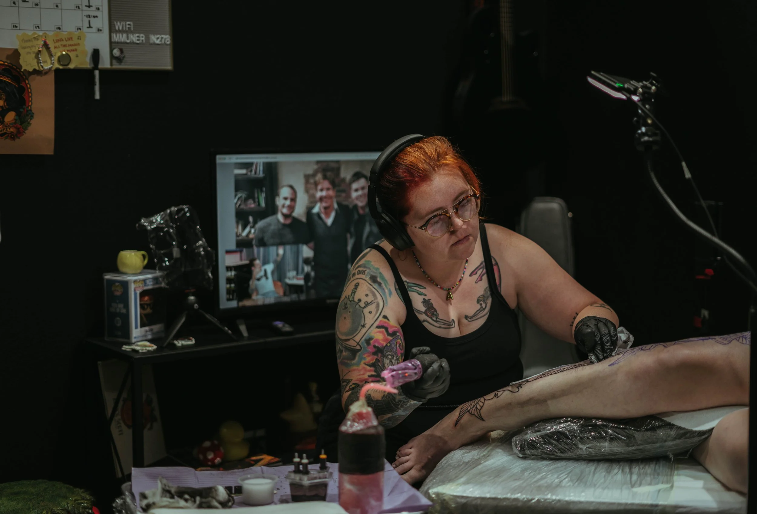 A woman with tattoos and glasses is tattooing a person's leg in a room with black walls. Behind her, a monitor displays a photo of three smiling men in a casual setting. The workspace has tattoo supplies, a computer, and a decorated table.