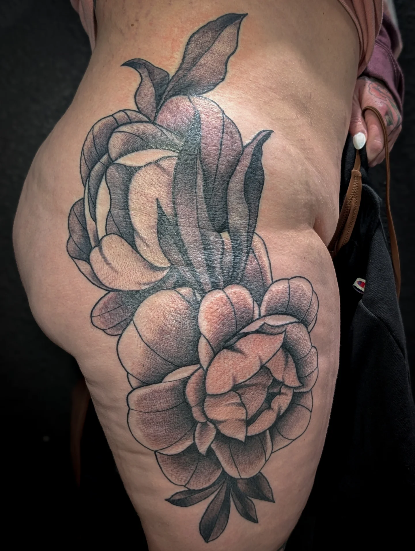 A detailed black and gray tattoo of a blooming flower with leaves on a person's upper thigh.