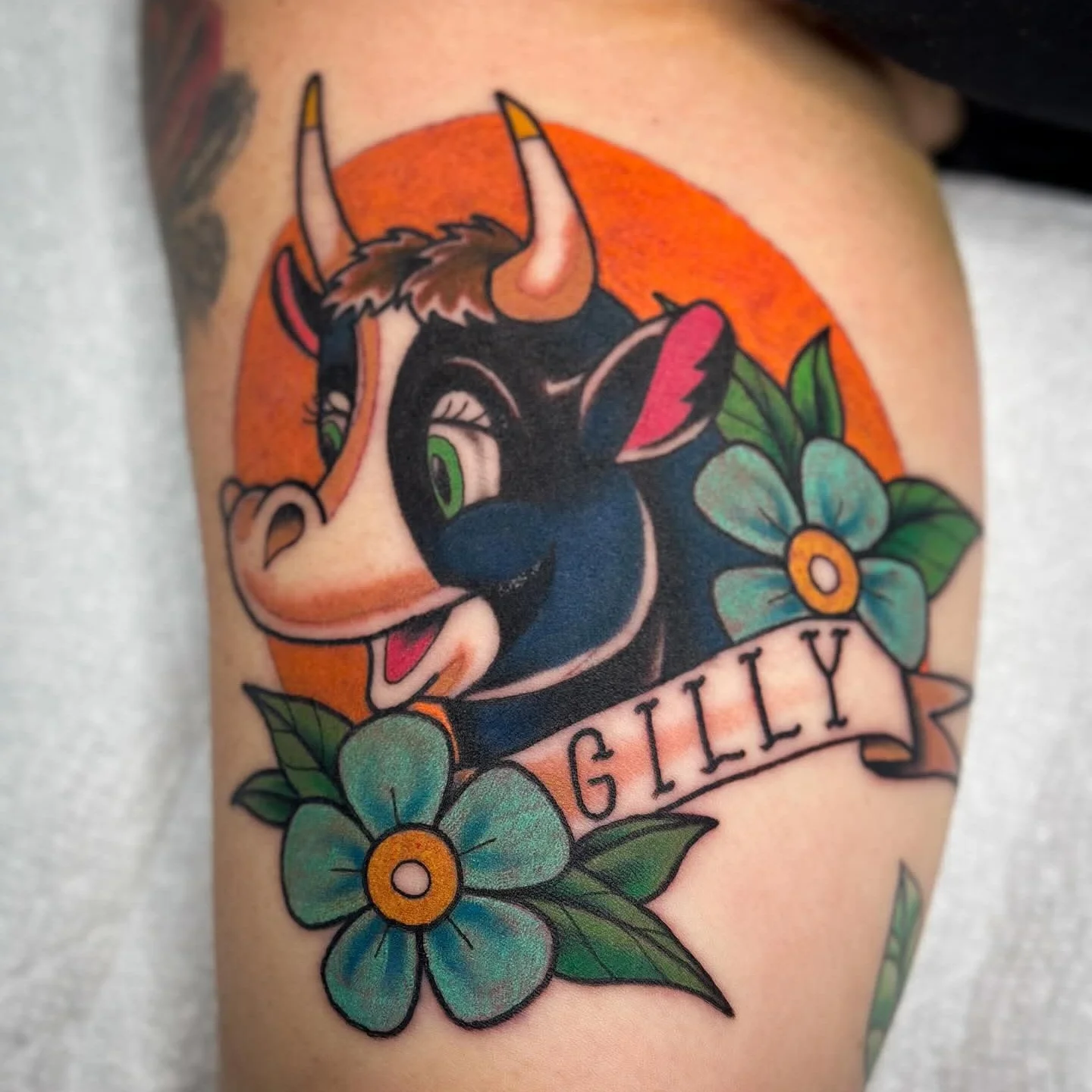 Colorful tattoo of a black cat with green eyes, surrounded by blue flowers and green leaves, with an orange background and a banner that reads 'GILLY'.