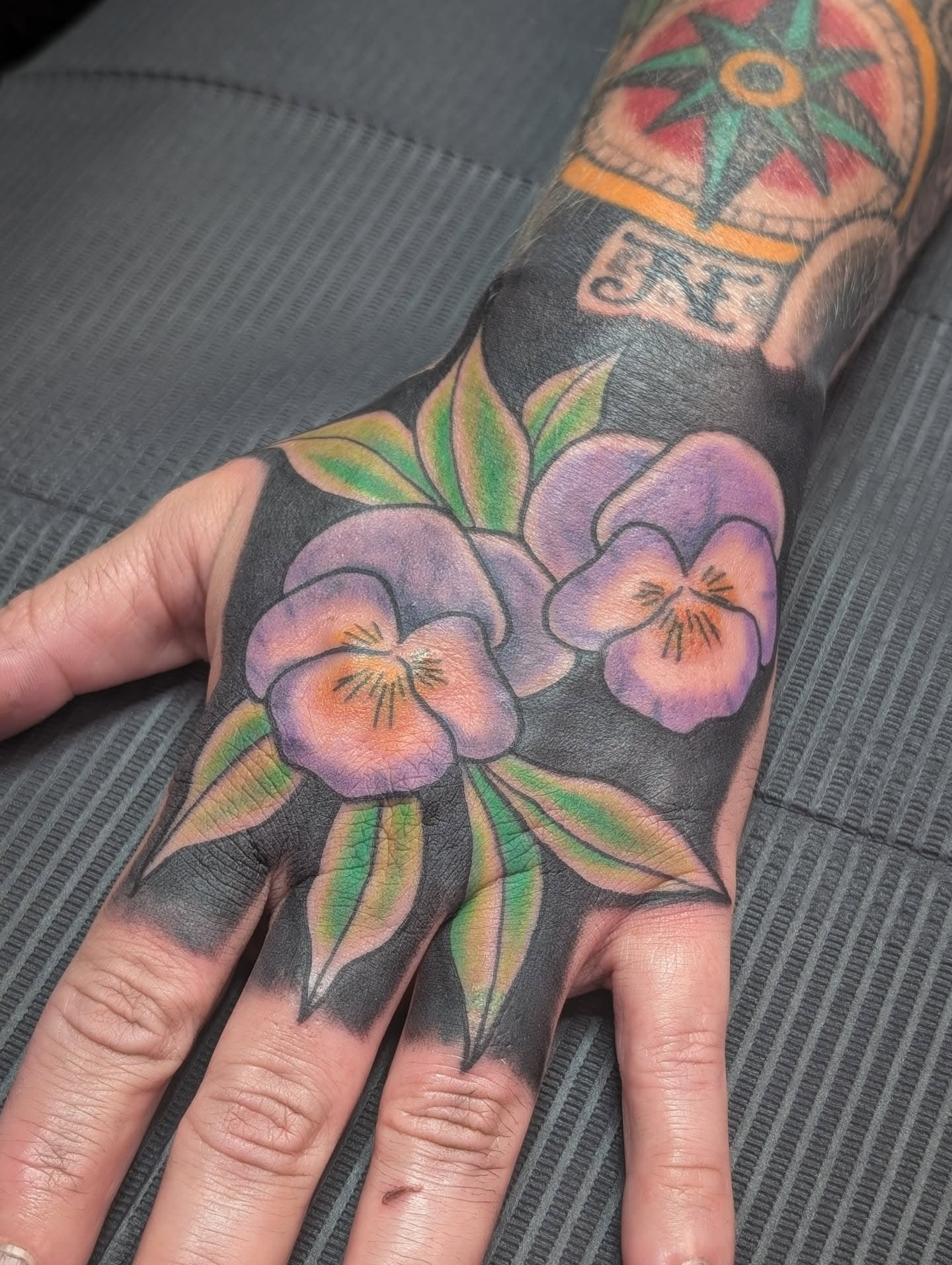 A tattoo of purple and orange flowers with green leaves on a person's arm. There is also a partial tattoo of a colorful star and the word that appears to be 'ART'.