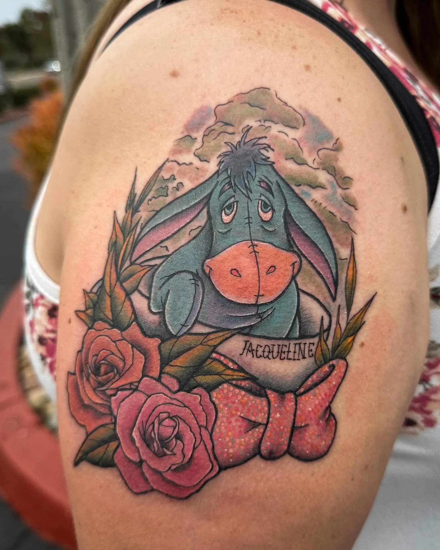 Tattoo of Eeyore from Winnie the Pooh with a background of clouds, roses, and the name Jacqueline beneath the character.