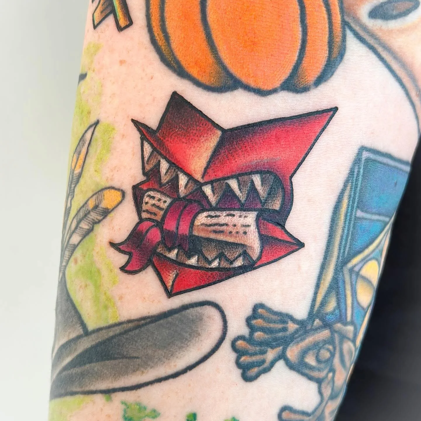 A tattoo of a cartoonish, open-mouthed monster with sharp teeth, red tongue, and spiked blue and red facial features.