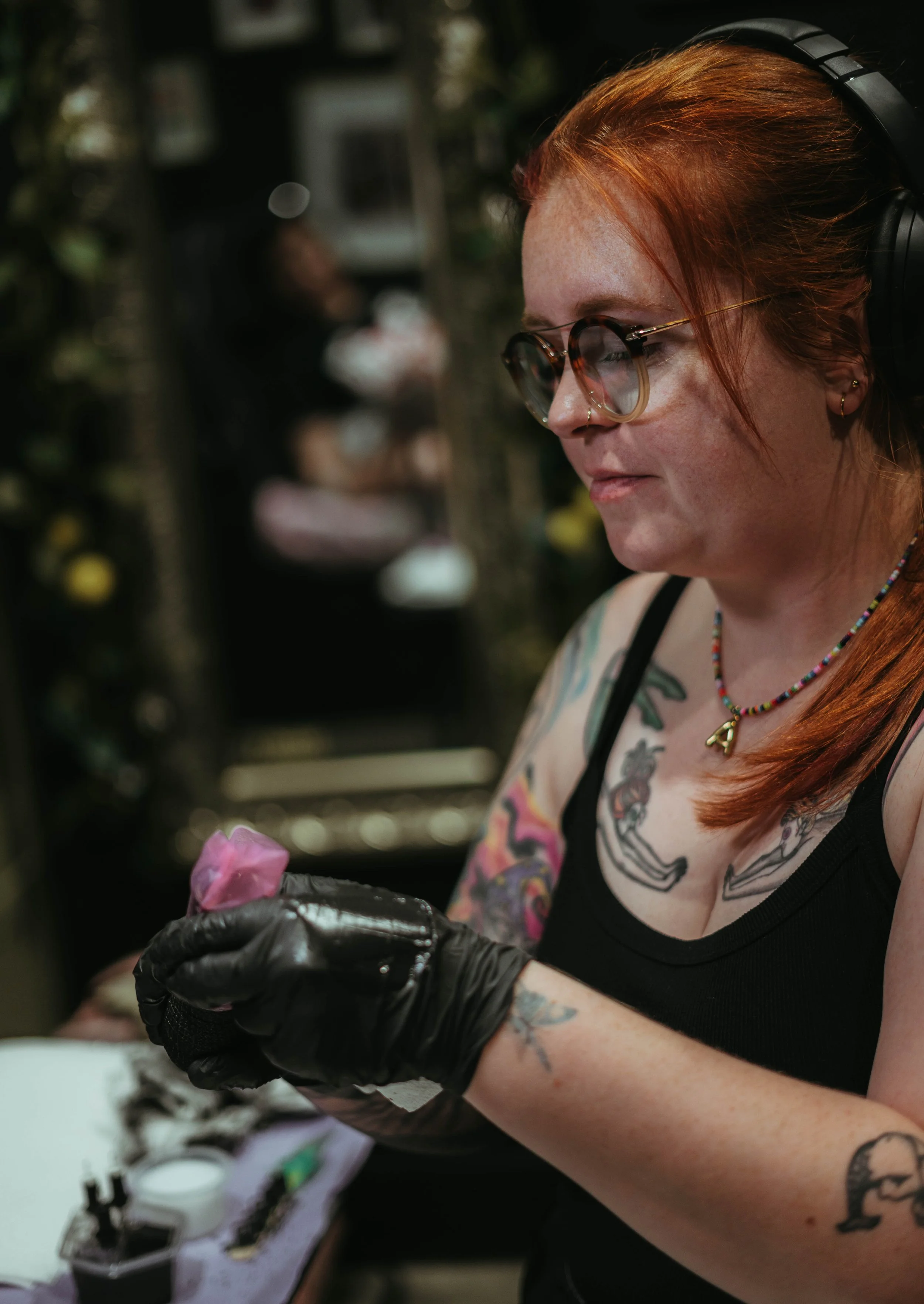 A woman with red hair, tattoos, glasses, and a nose piercing, wearing black gloves and headphones, tattooing on someone's arm in a tattoo studio.