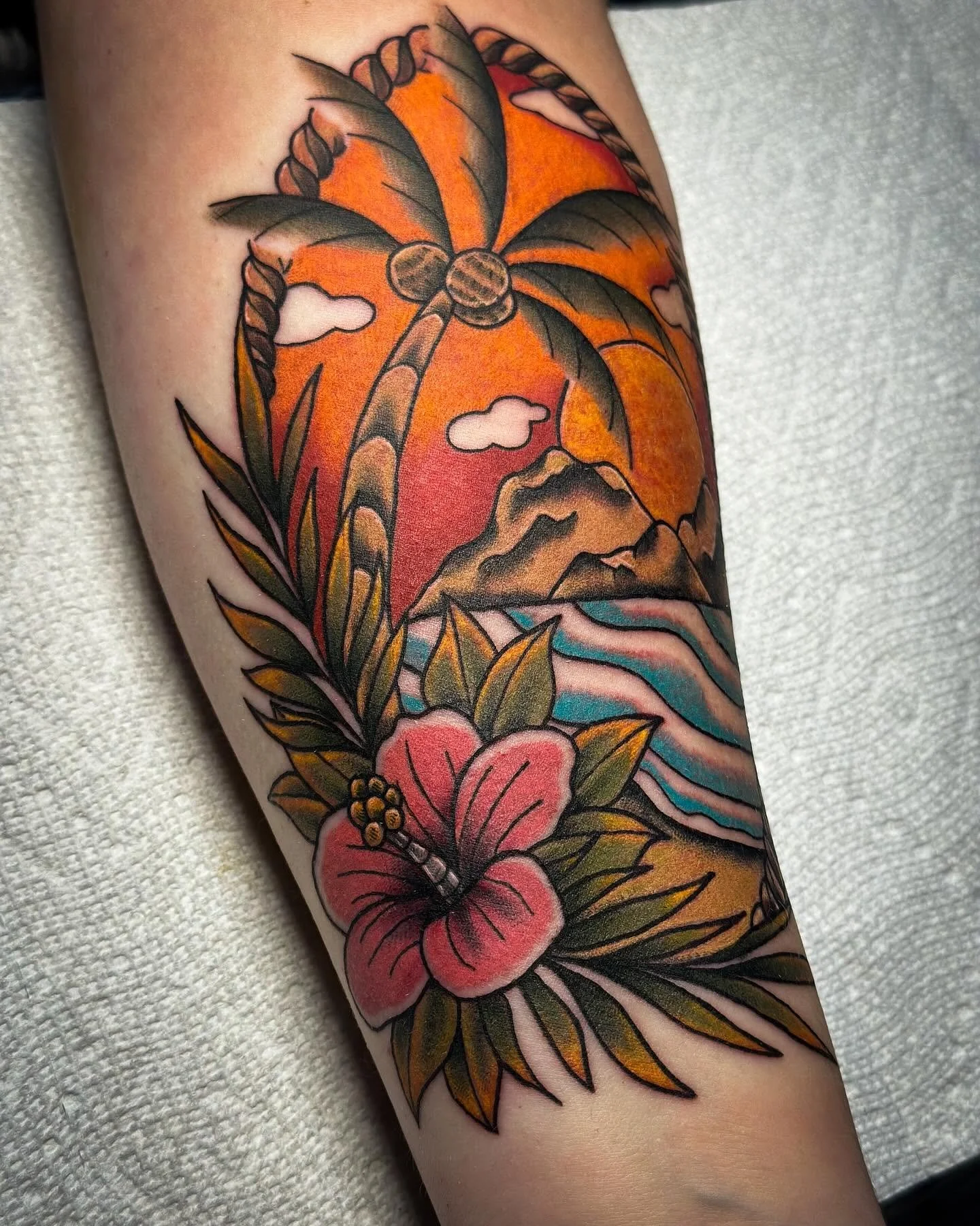 A colorful tattoo featuring a palm tree, mountains, a sunset, clouds, a hawaiian lei, and a hibiscus flower on a person's arm.