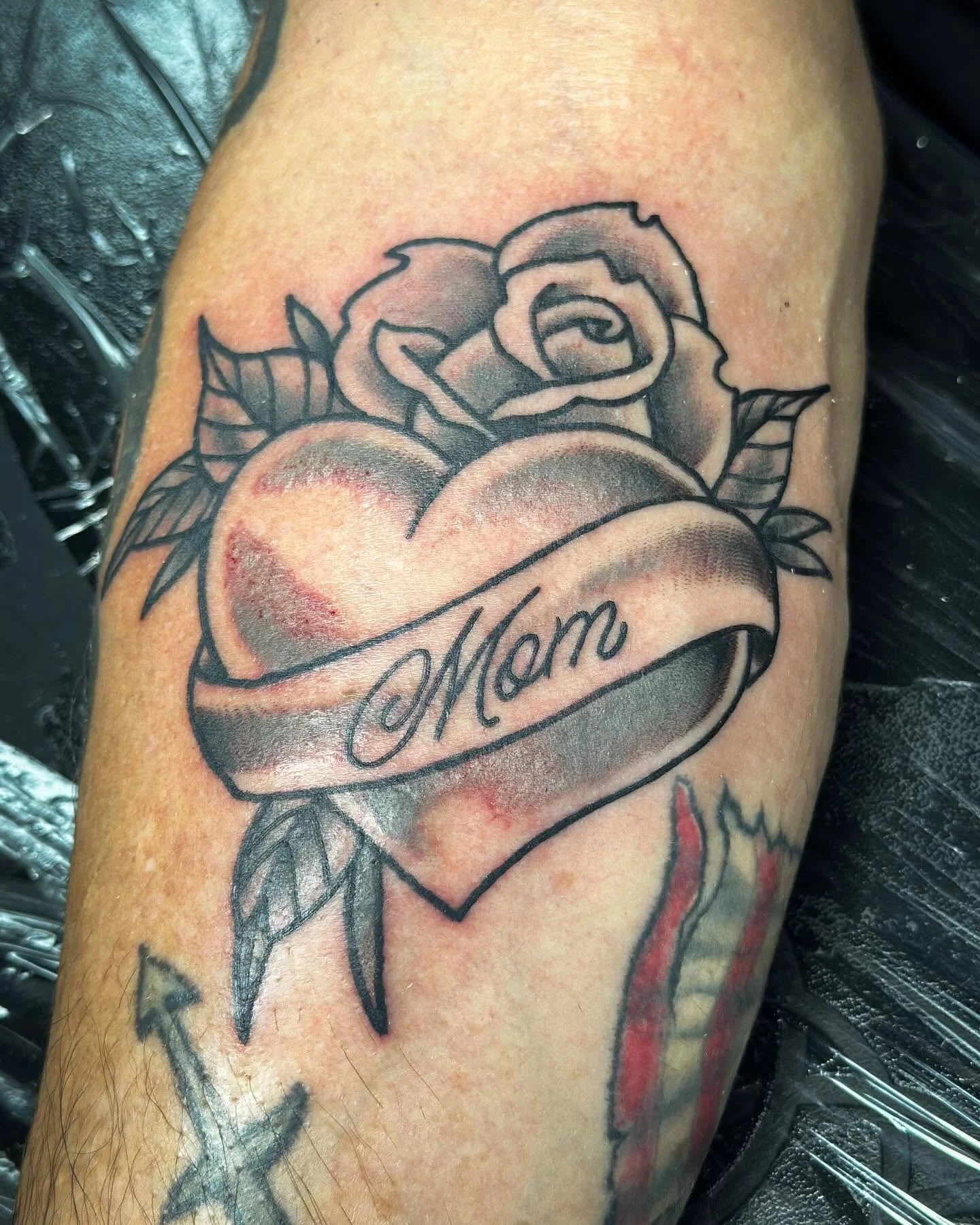 Tattoo of a heart with a ribbon banner saying 'Mom' and a rose placed on top of the heart.
