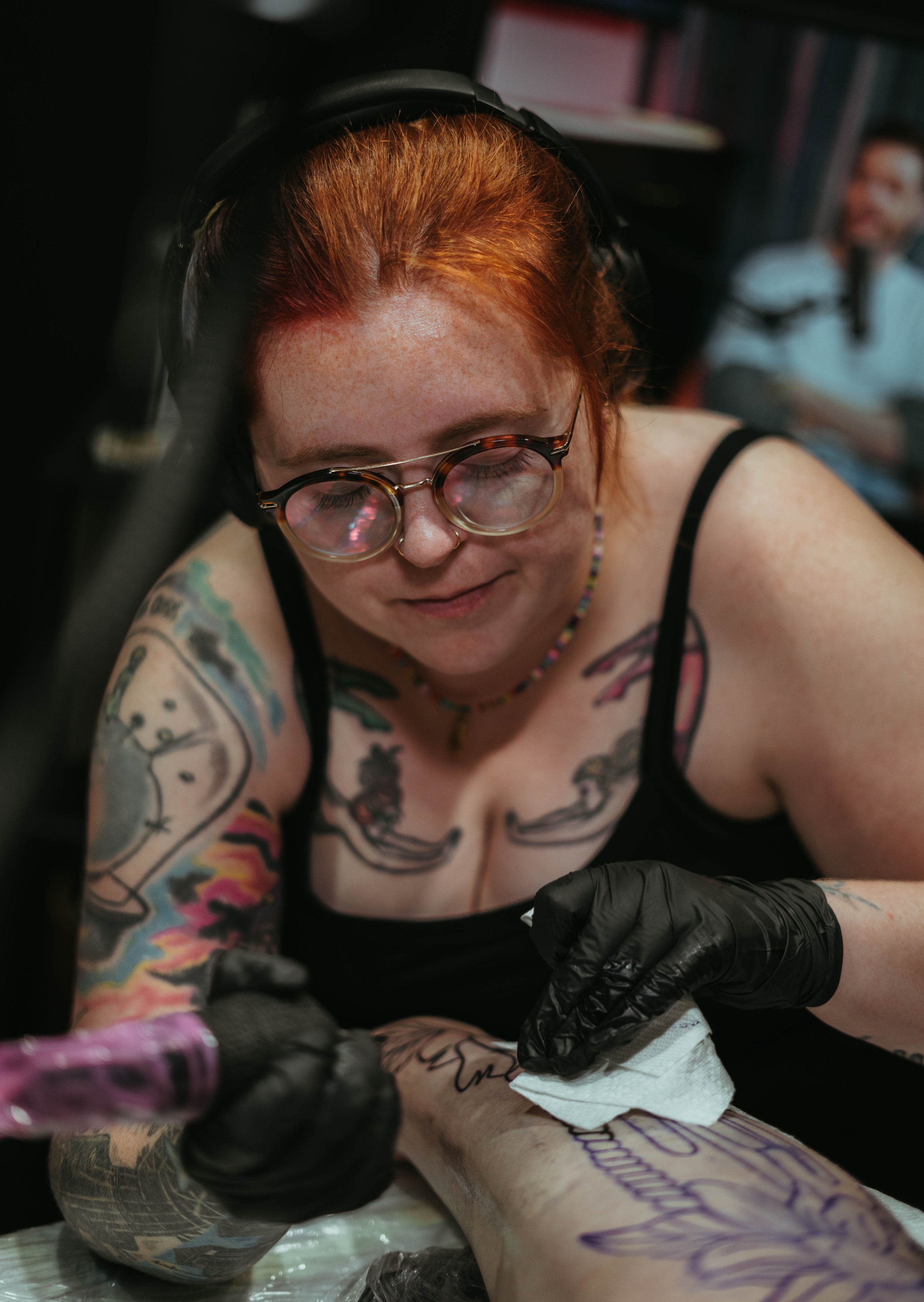 A person with tattoos on their arms and chest, wearing glasses and headphones, is getting a tattoo applied on their leg by a tattoo artist wearing black gloves.