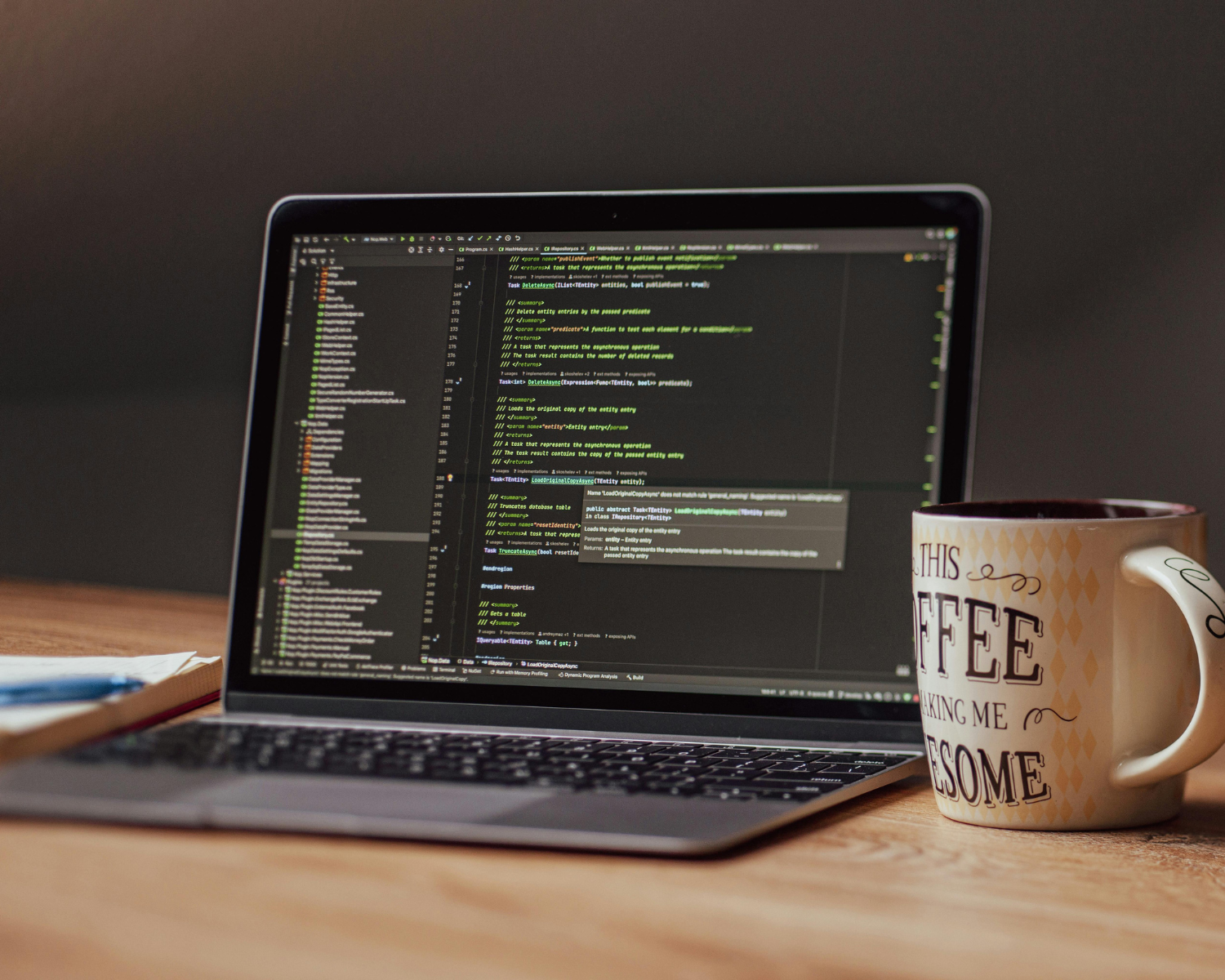 A laptop computer on a wooden desk displaying lines of programming code in a dark-themed editor. To the right of the laptop, there is a coffee mug with the words "This coffee is making me awesome" written on it. A closed notebook with a pen is partially visible on the left side of the image.