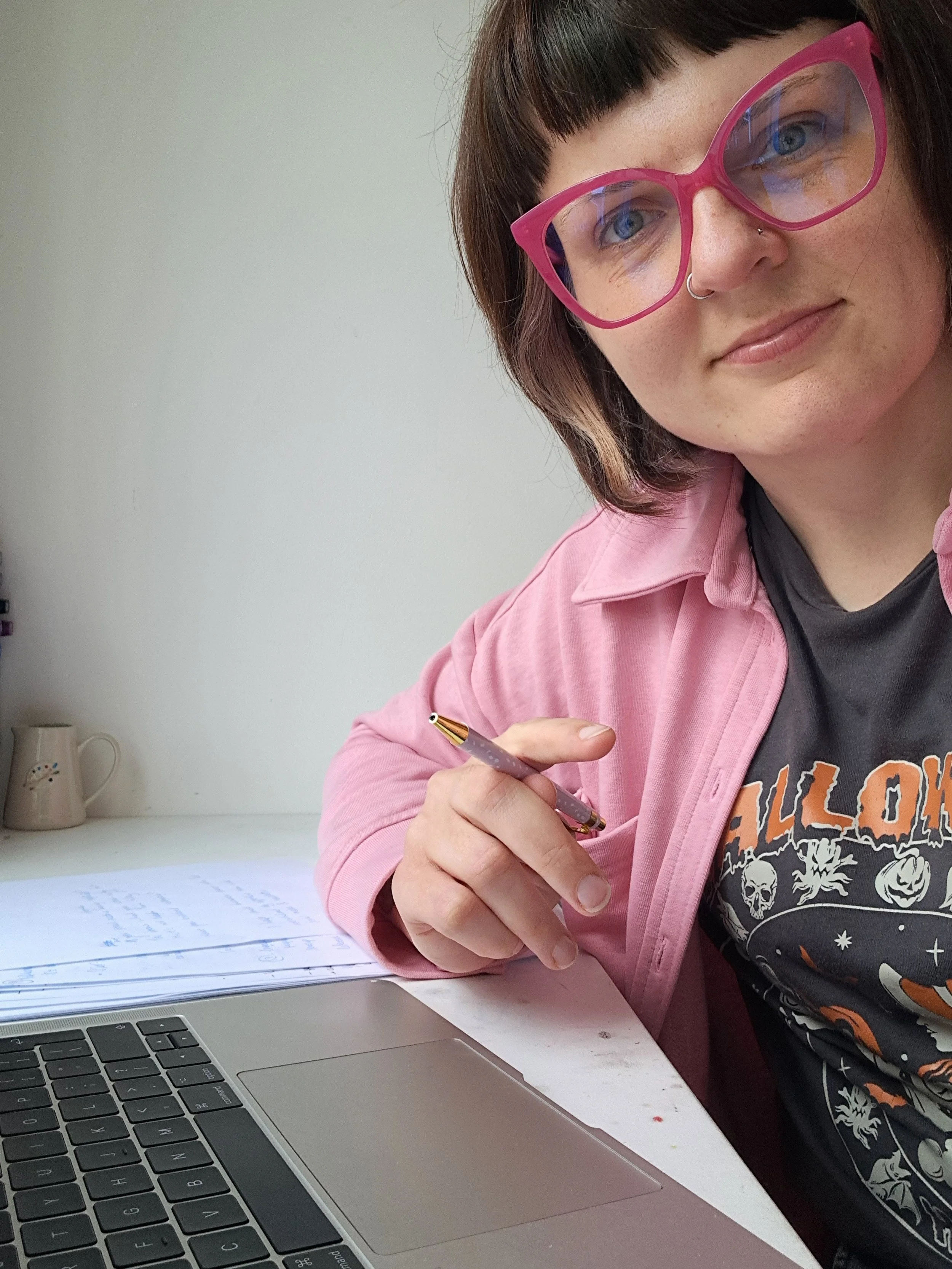 A woman with short dark hair, pink glasses, and a nose ring, wearing a pink jacket and a Halloween-themed t-shirt, sitting at a desk with a laptop, holding a pen over papers with handwritten notes, in front of a plain light-colored wall.