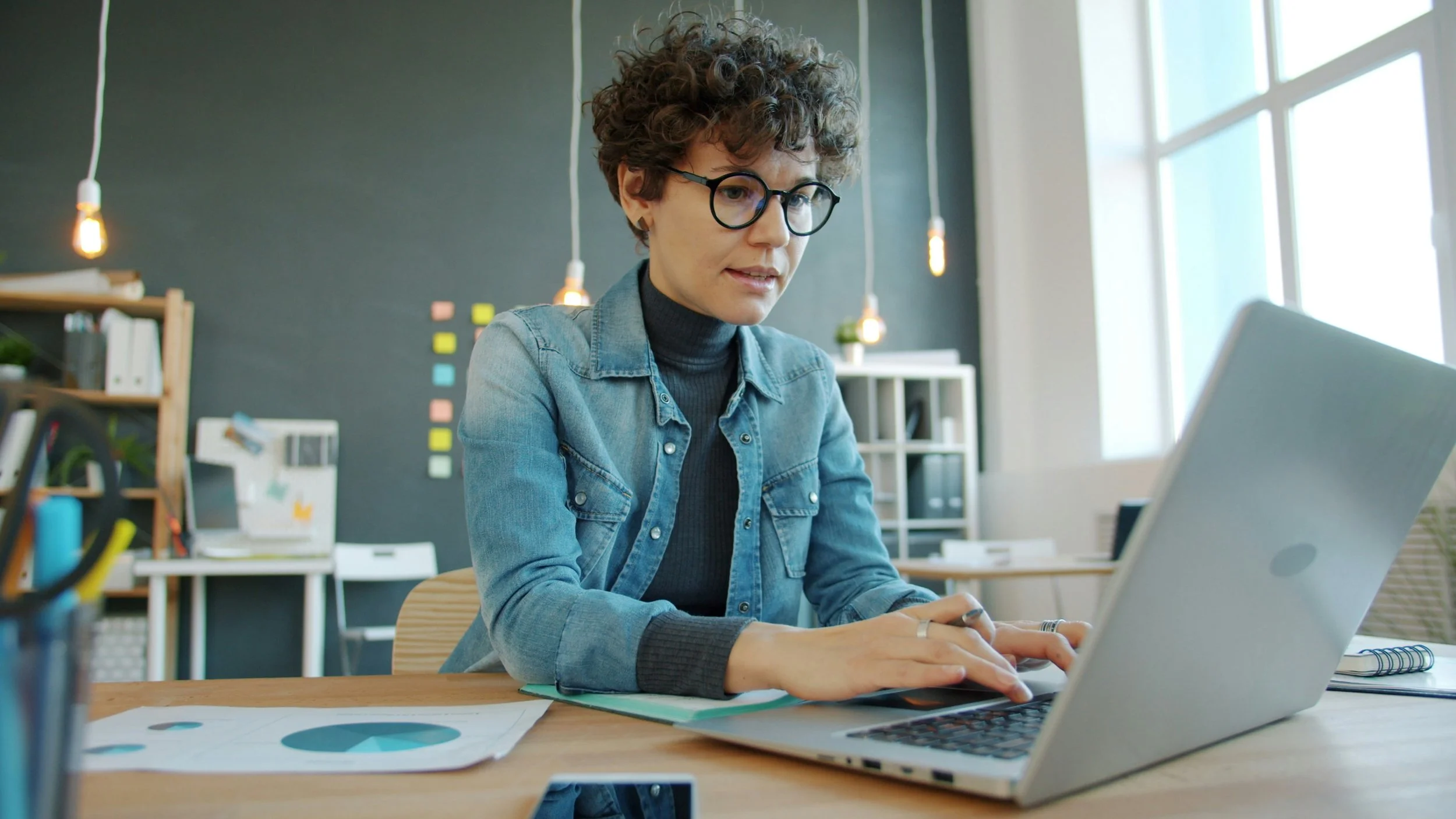 Lady with black round glasses sitting at laptop and typing a blog in her office room with pens and post it notes.