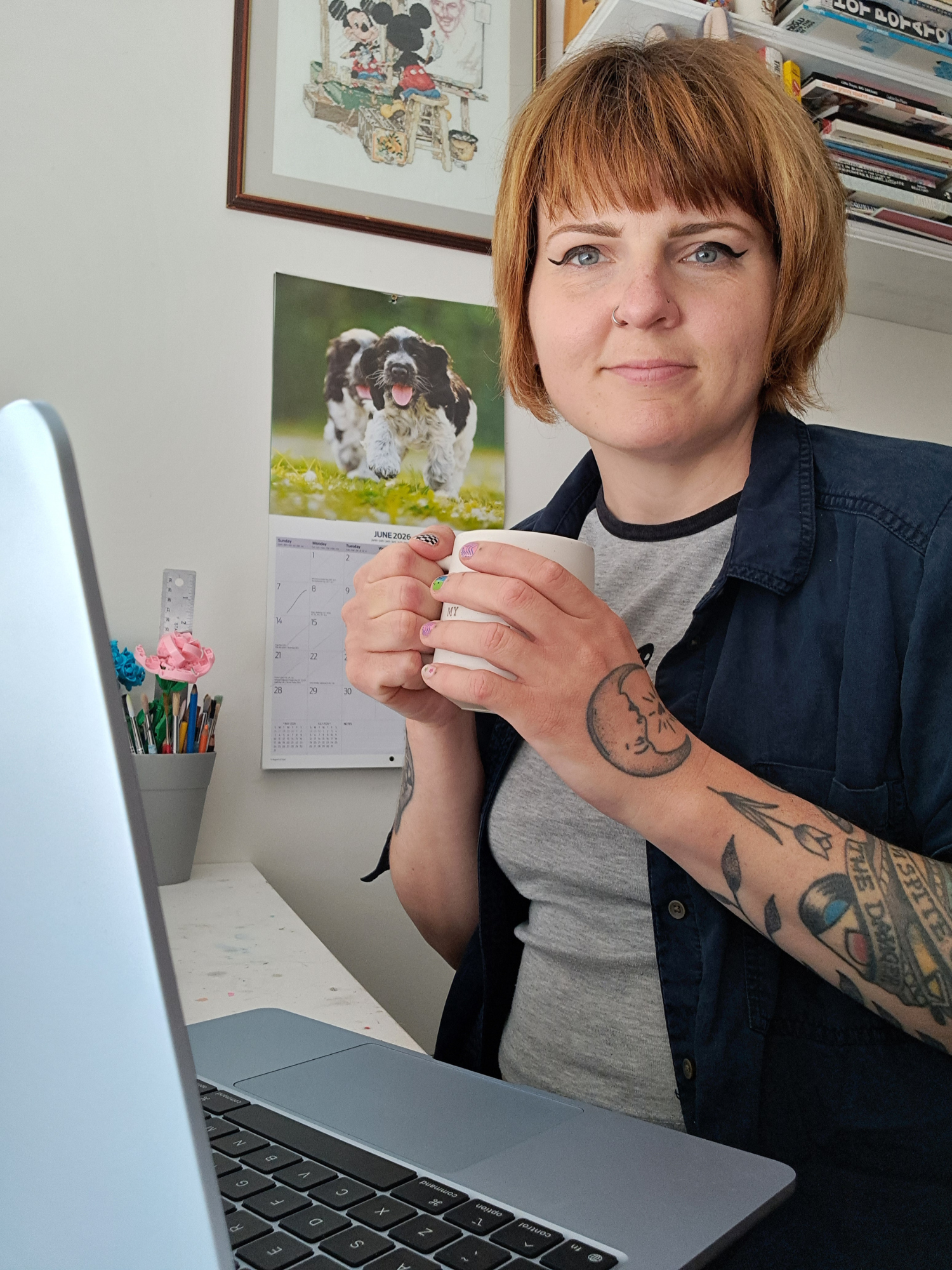 Lady with copper hair and blue eyes is smiling at the camera and sitting at her laptop with a calendar in the background