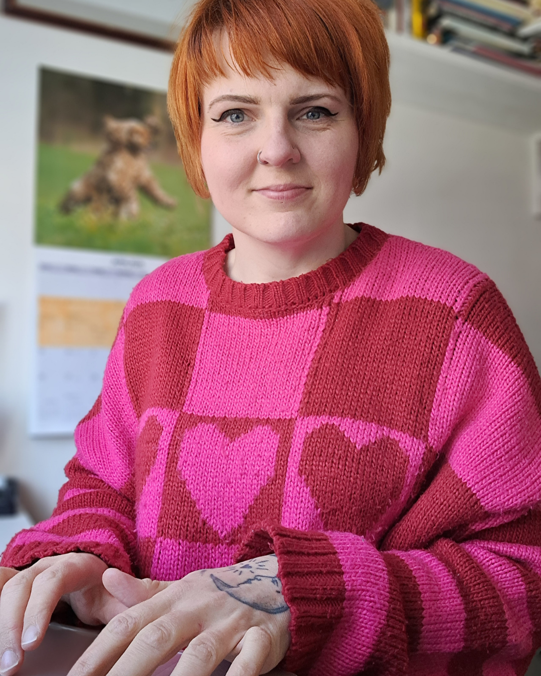 A woman with short red hair, wearing a pink and red checkered sweater with heart patterns, sitting at a desk with a computer. There is a calendar and a poster of a dog in the background.